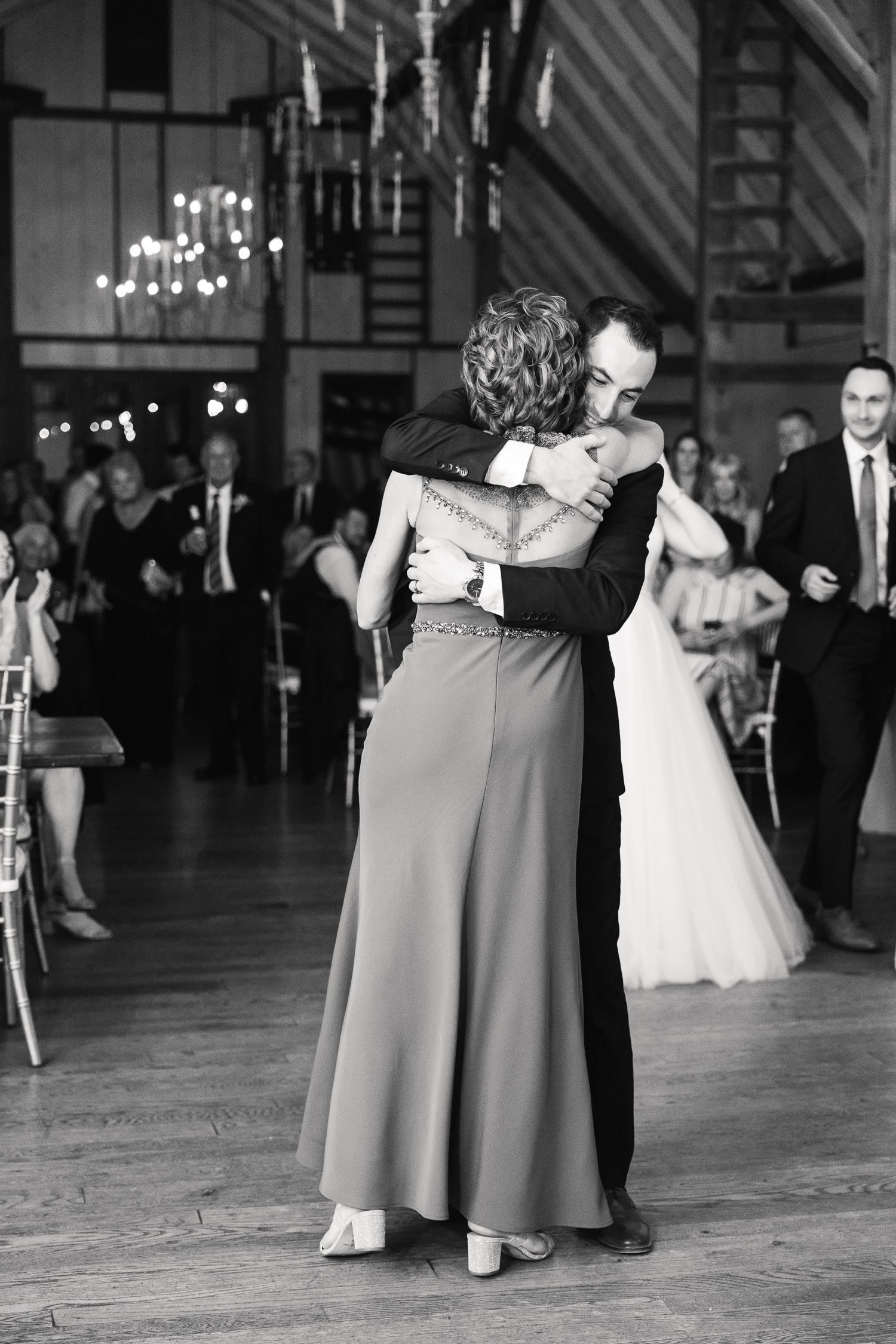 A black and white photo of two dancers embracing on a reception dance floor under chandeliers.
