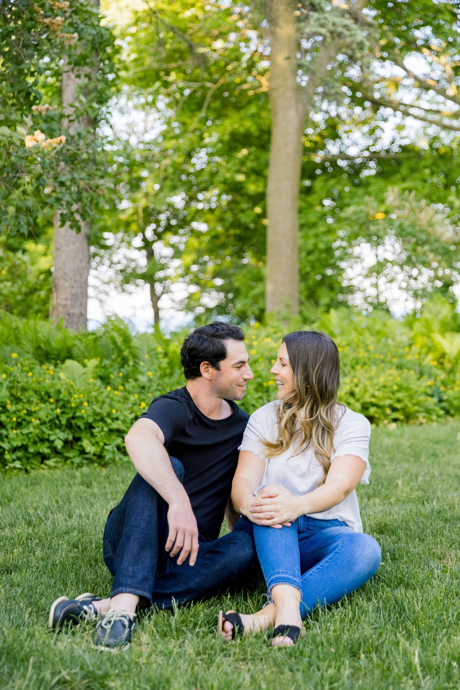 A couple sits together on grass with tall trees creating a natural backdrop.