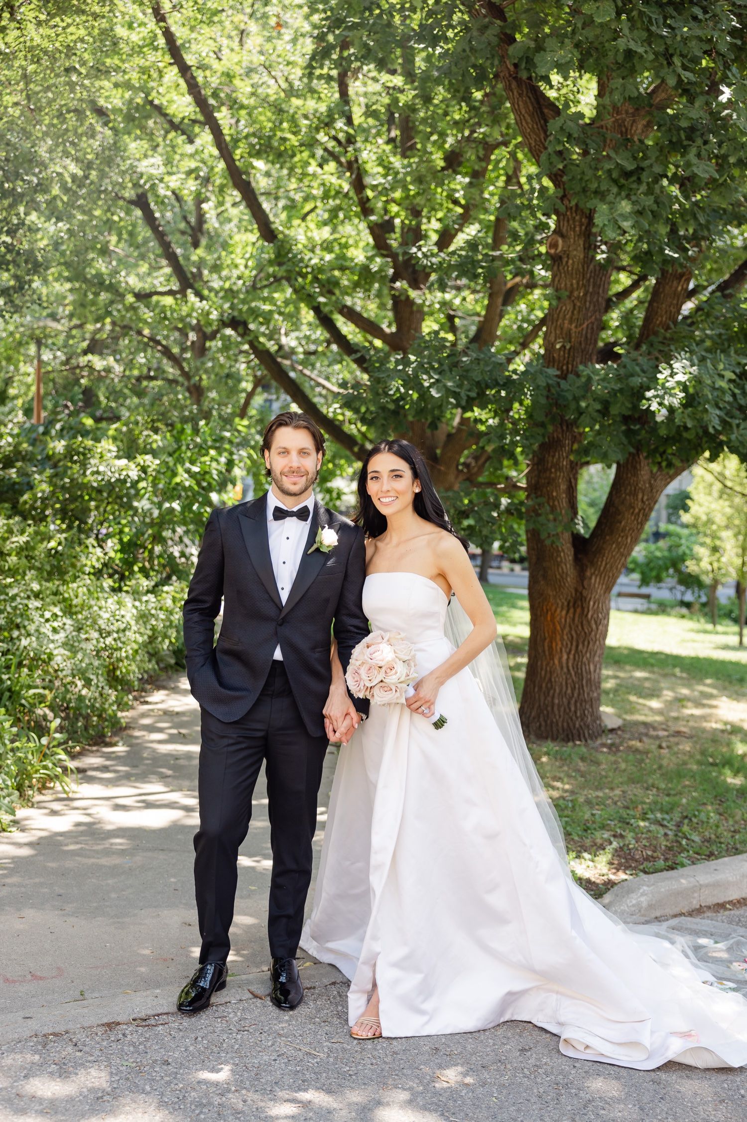 Elegant wedding couple poses on pathway under lush green trees in summer garden setting.