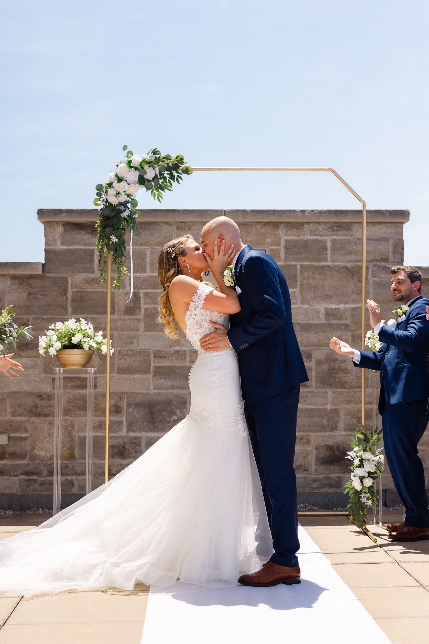First kiss as newlyweds under a modern geometric arch decorated with white flowers and greenery.