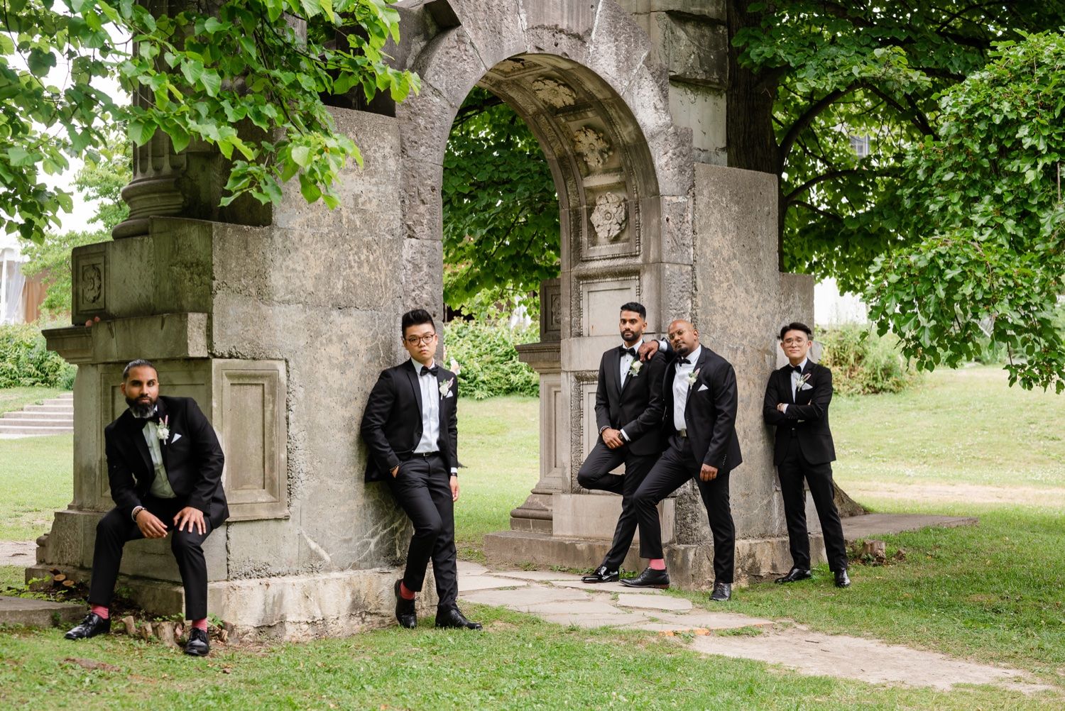 Groomsmen in black tuxedos posing casually around stone archway structure.