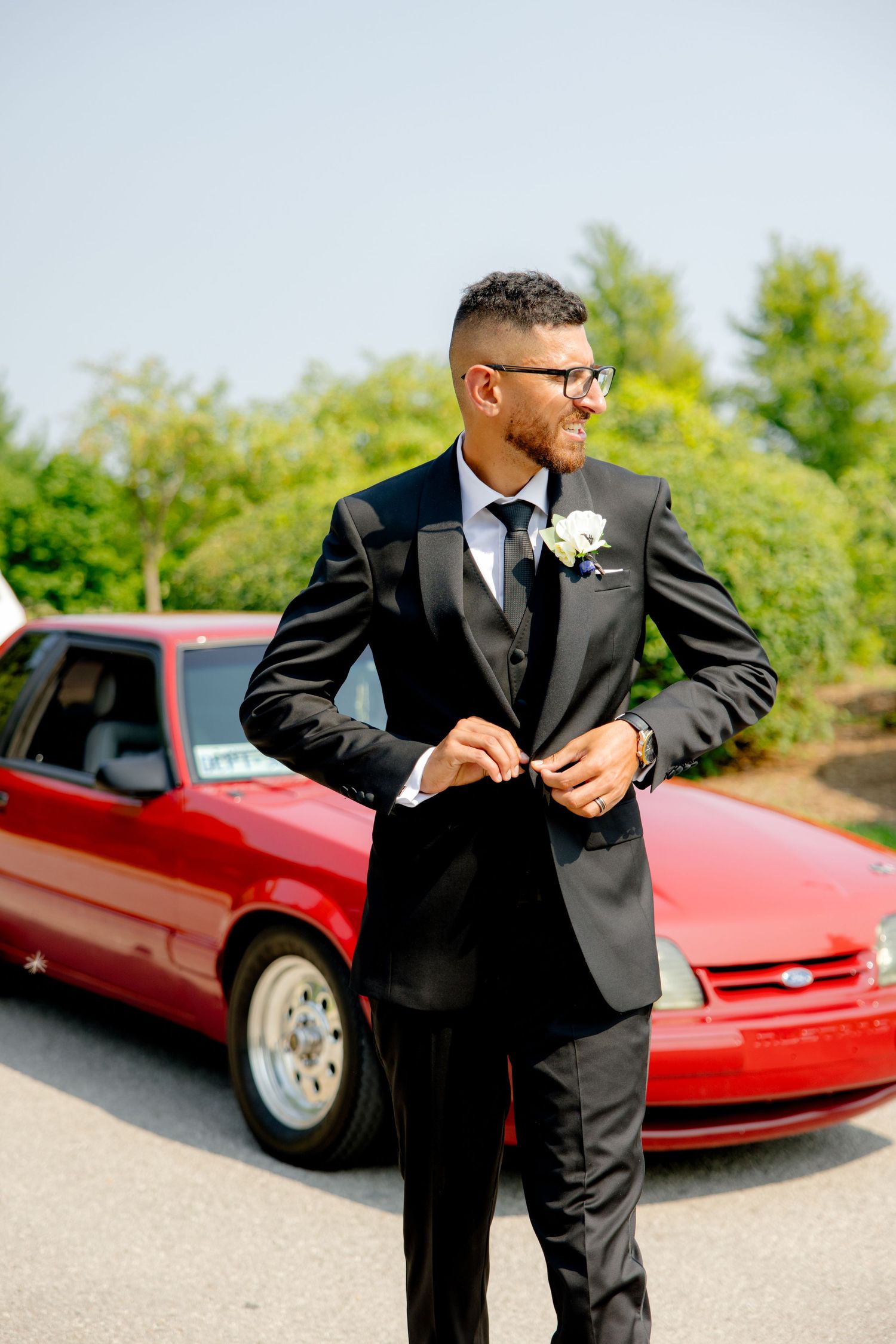 Groom adjusts his suit jacket while standing next to a red classic car outdoors.