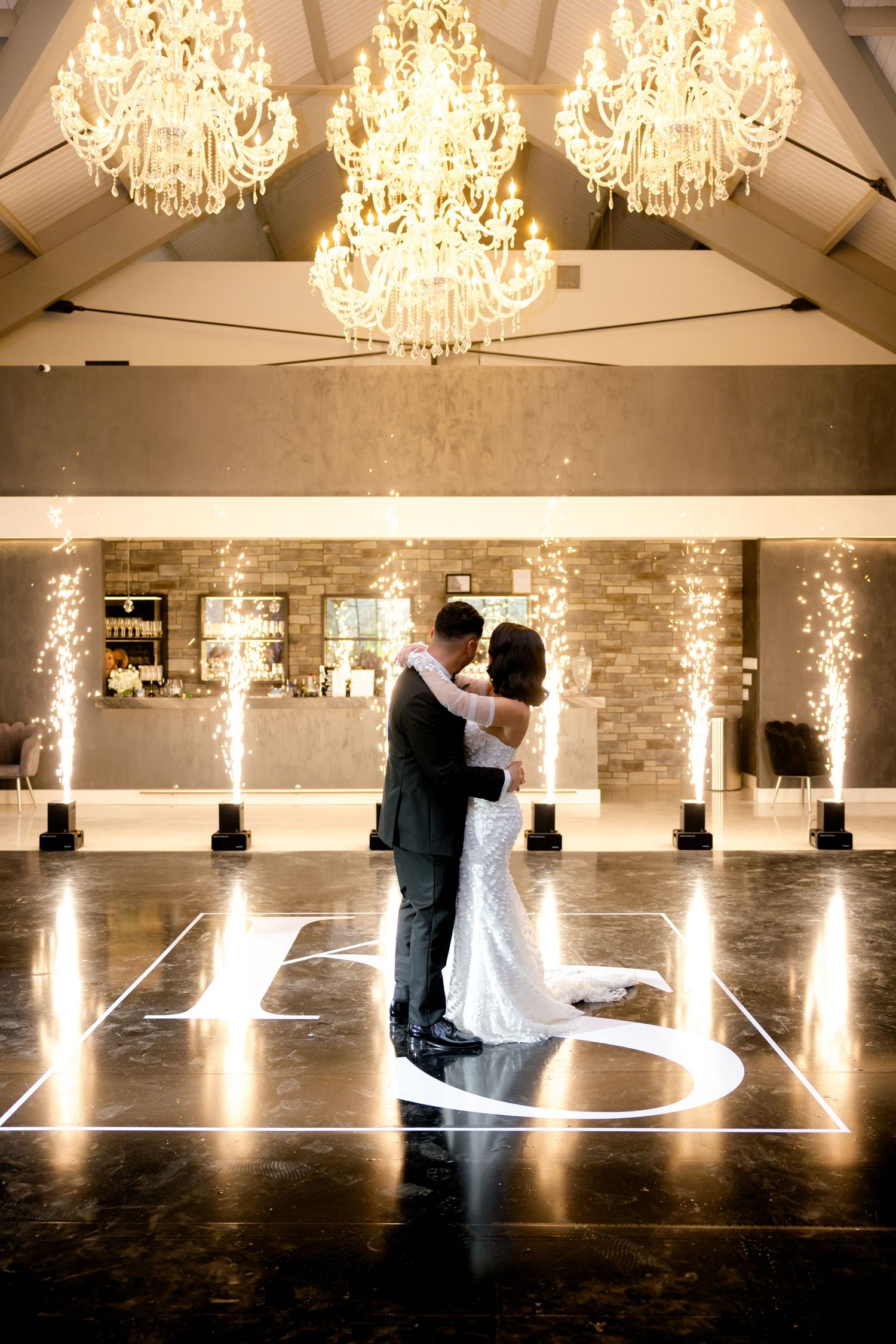 Newlyweds embrace on illuminated dance floor under ornate crystal chandeliers.