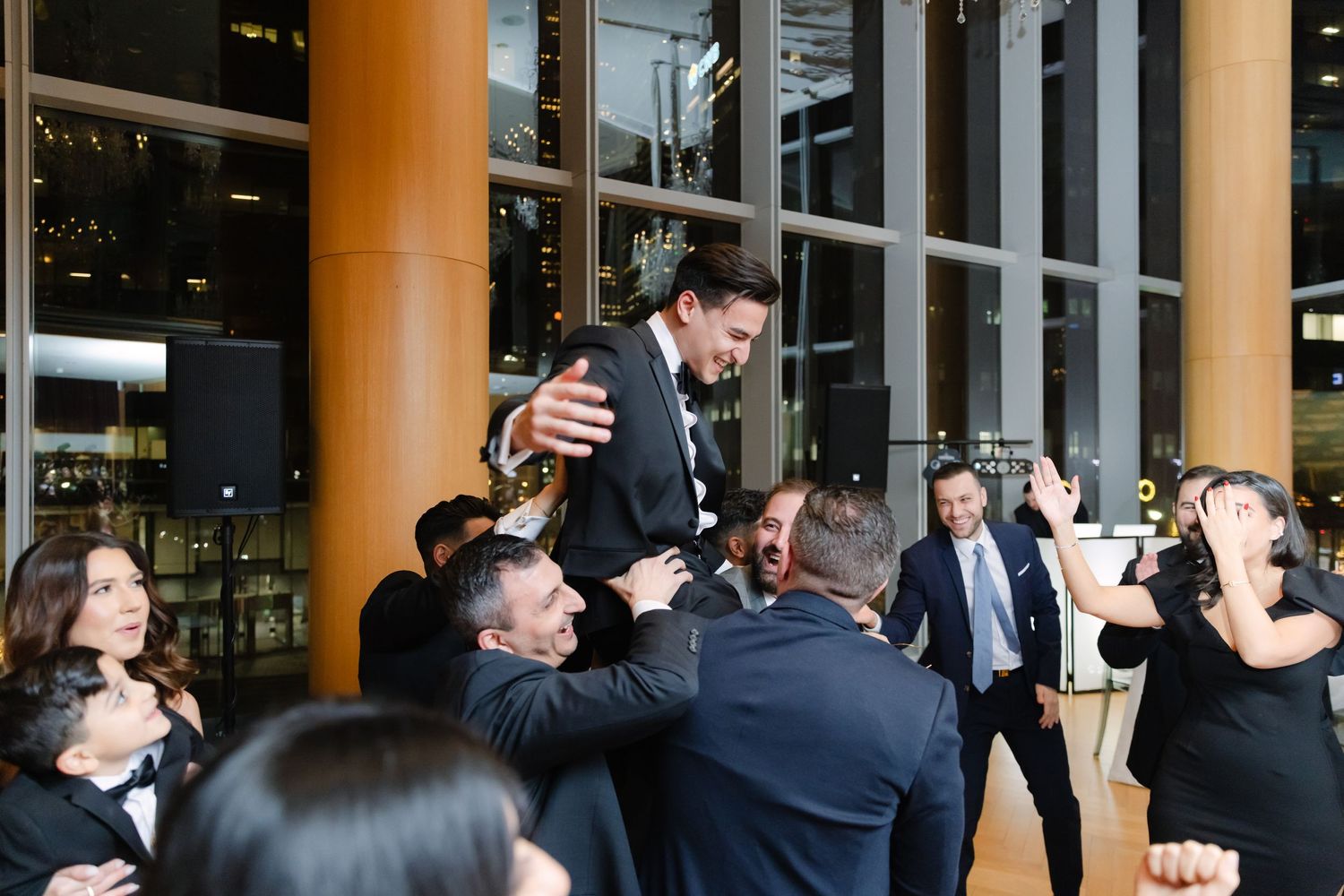 Wedding guests lift the groom on shoulders during traditional celebration dance at indoor venue.