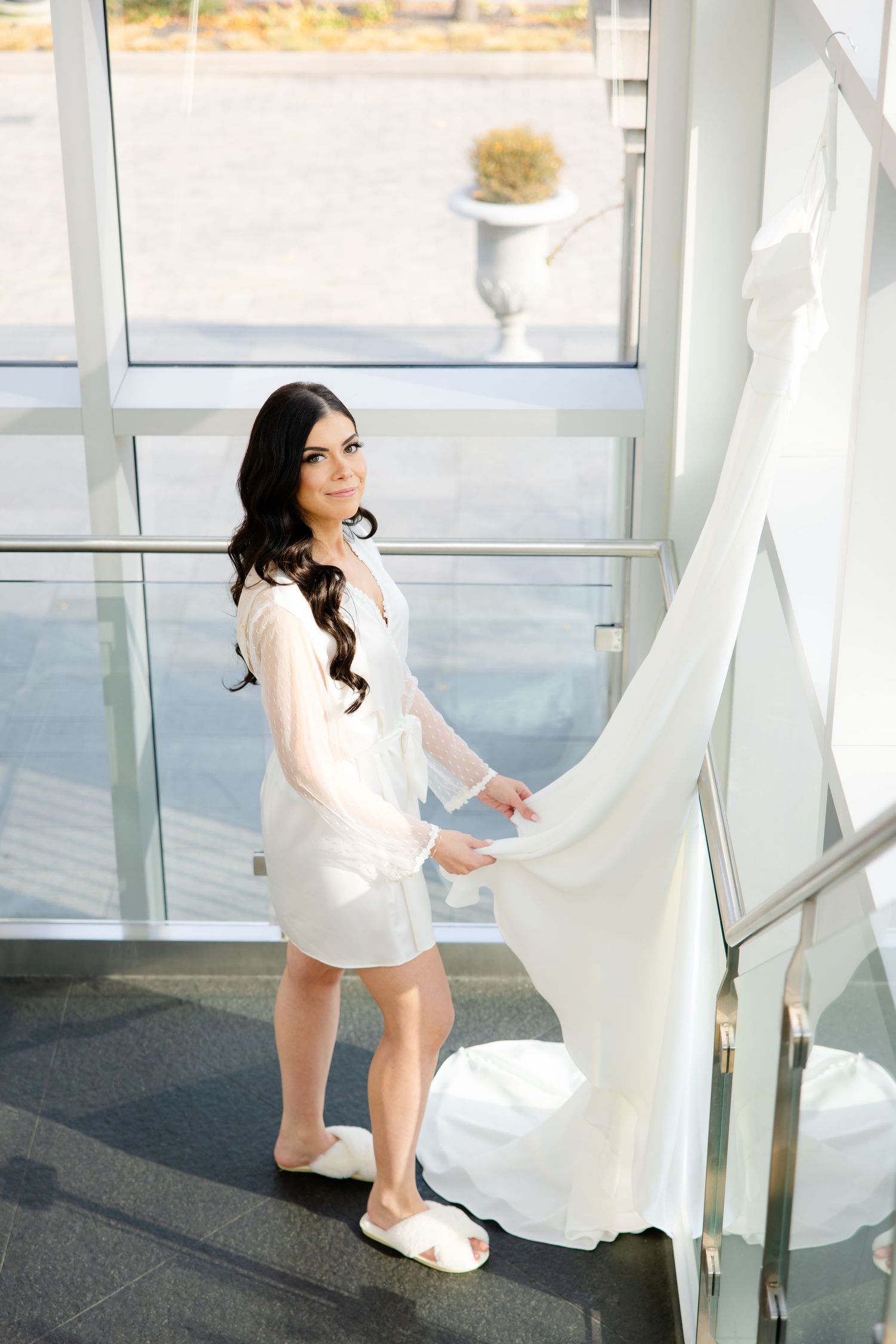 A bride in a short white robe and slippers stands by a window in preparation for her wedding day.