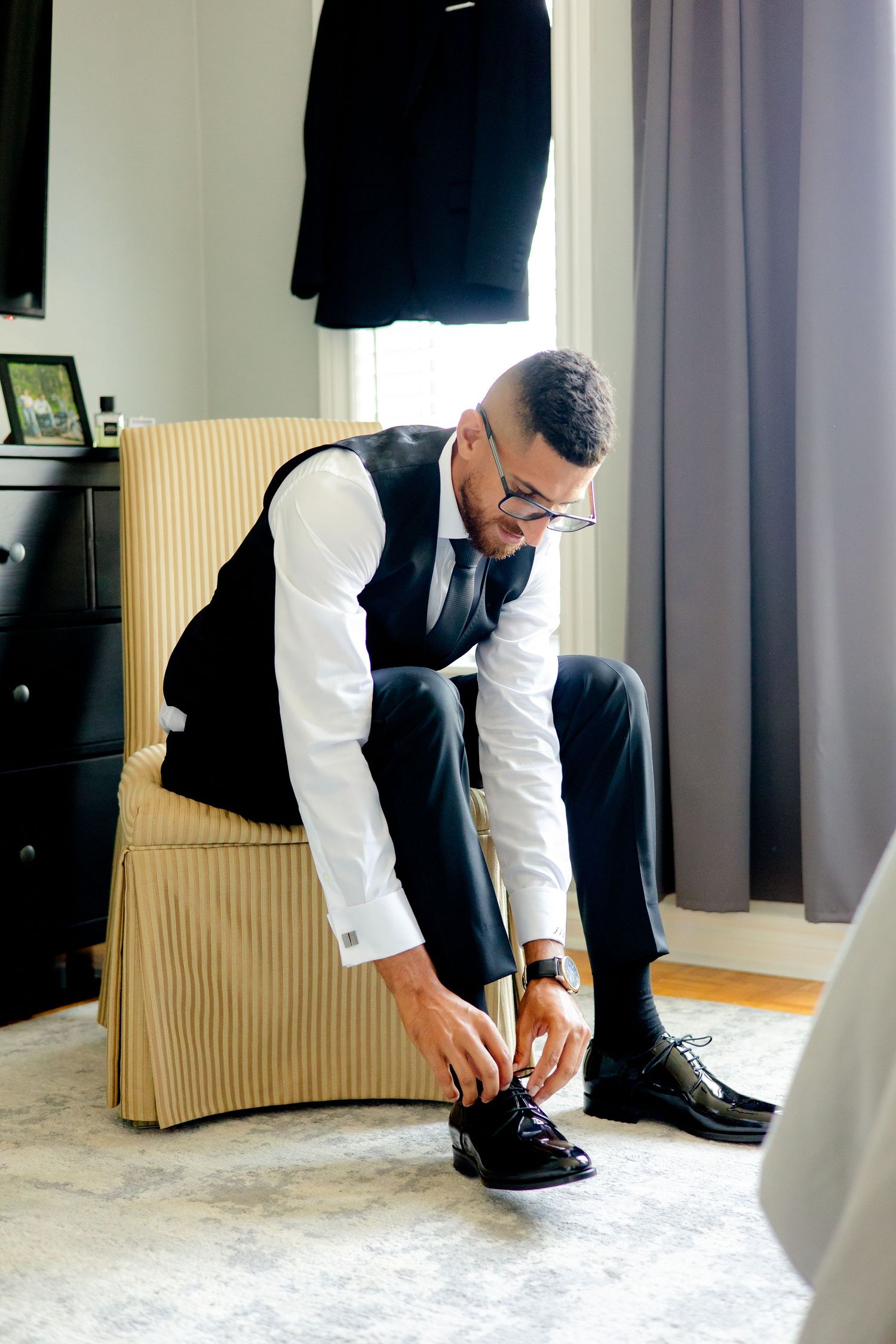 Well-dressed person in a vest and dress shirt laces up formal black shoes while seated.