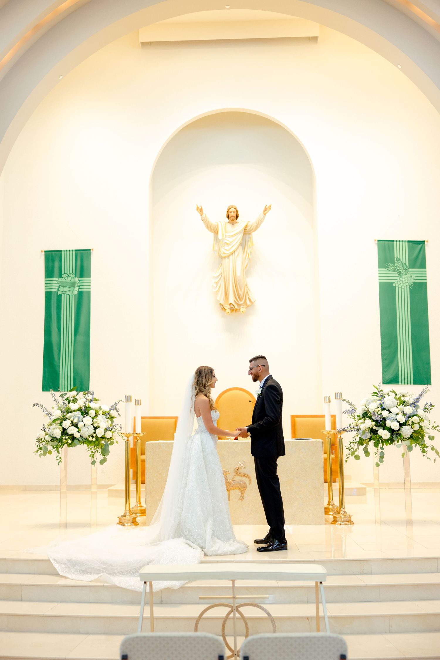 A ceremony moment captured beneath a golden Jesus statue with elegant white floral arrangements.