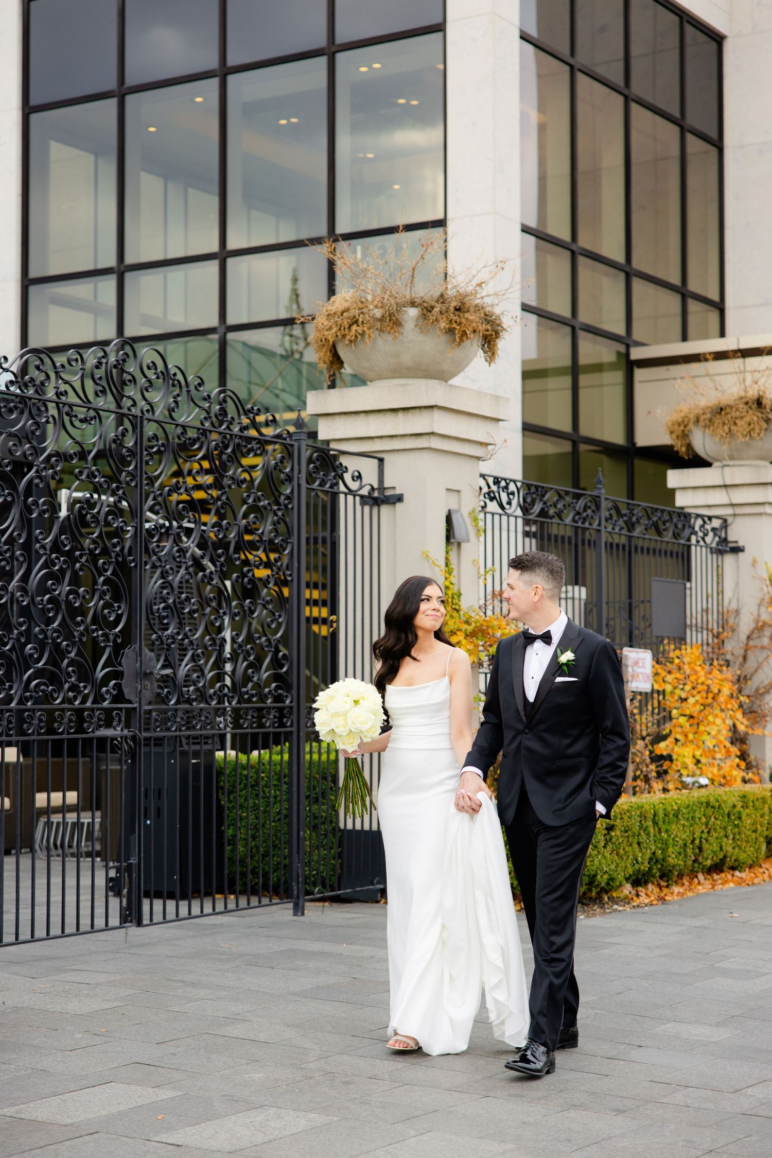 Elegant newlyweds pose outside a modern building with large windows and ornate iron gates.