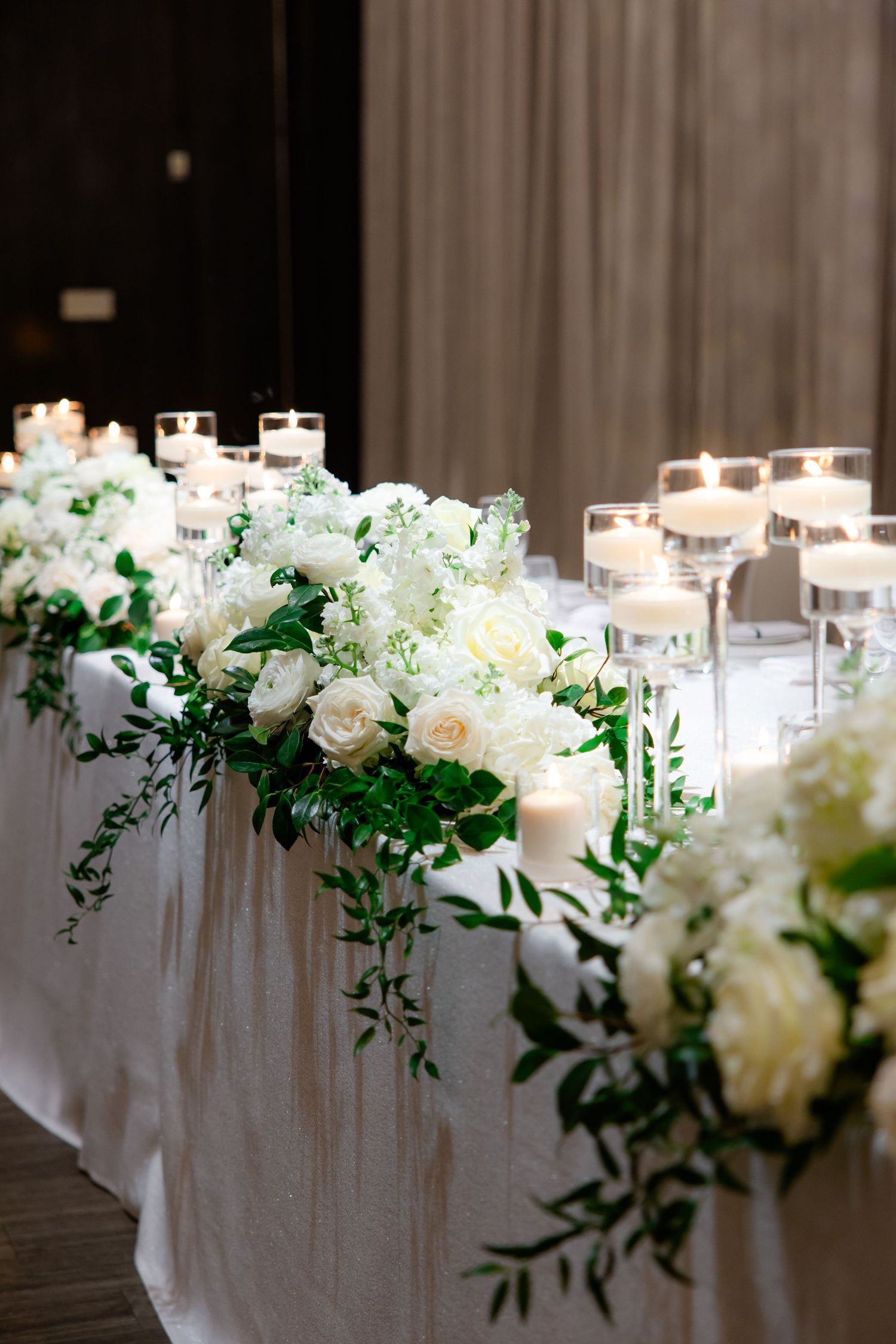 White floral table decorations with glowing candles along wedding reception tablecloth.