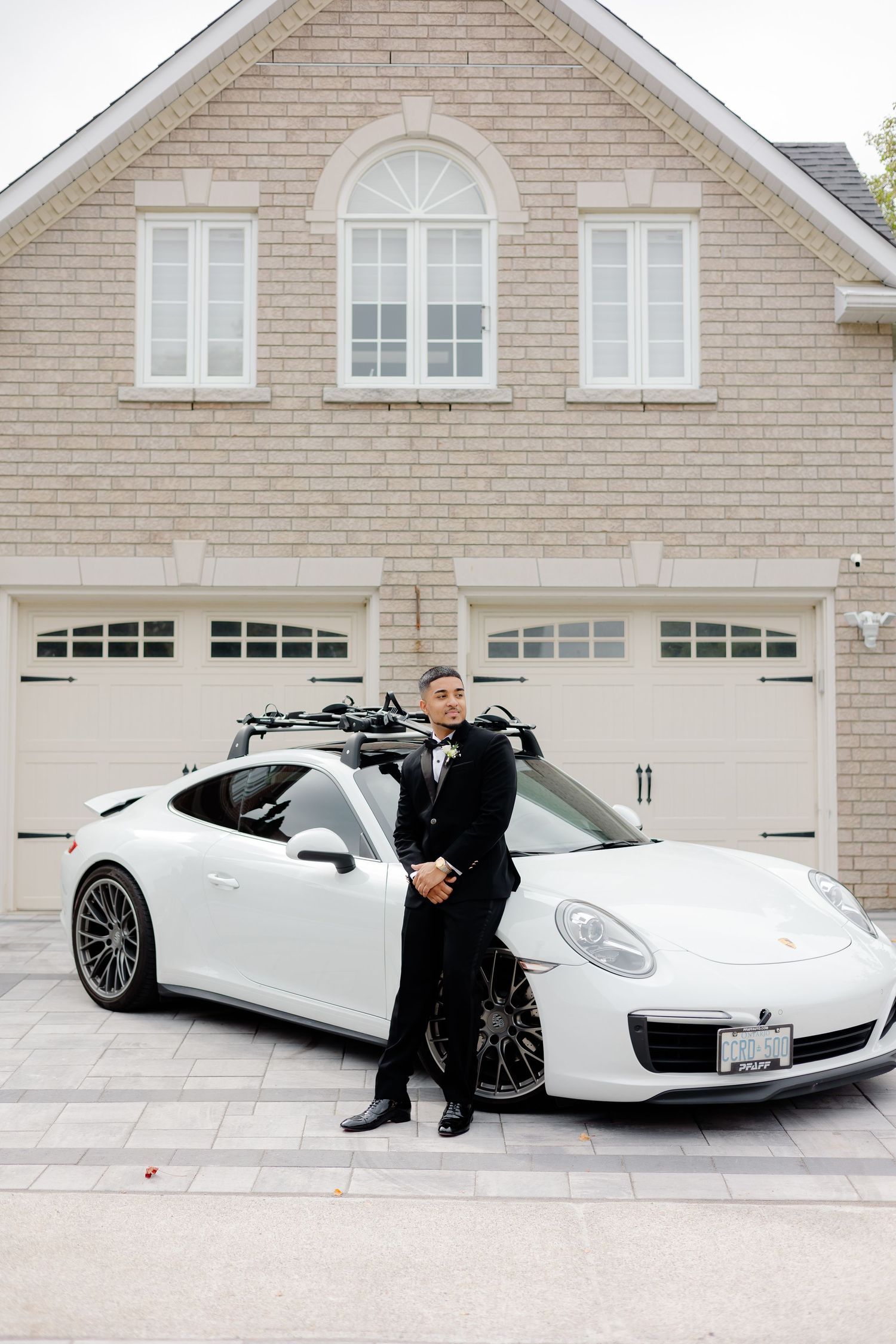 A person in formal attire stands beside a white Porsche in front of a modern brick house.
