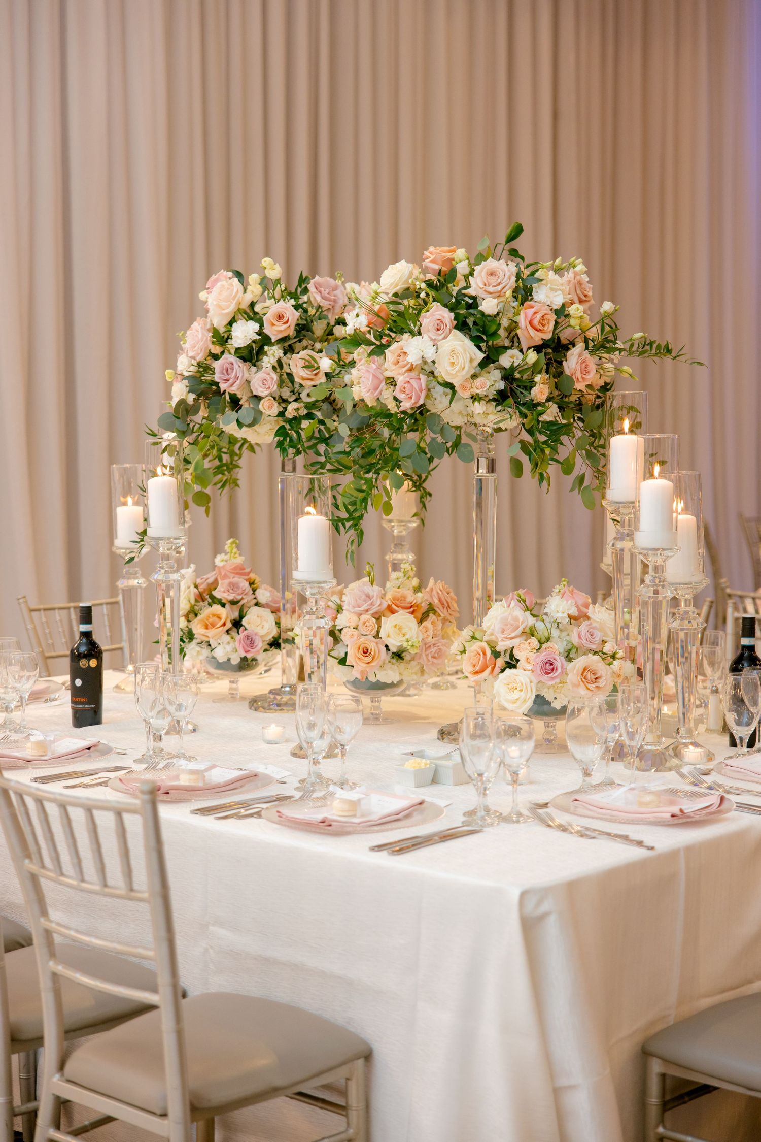 Elevated floral centerpiece with blush roses and greenery on white tablecloth with glass candle holders.
