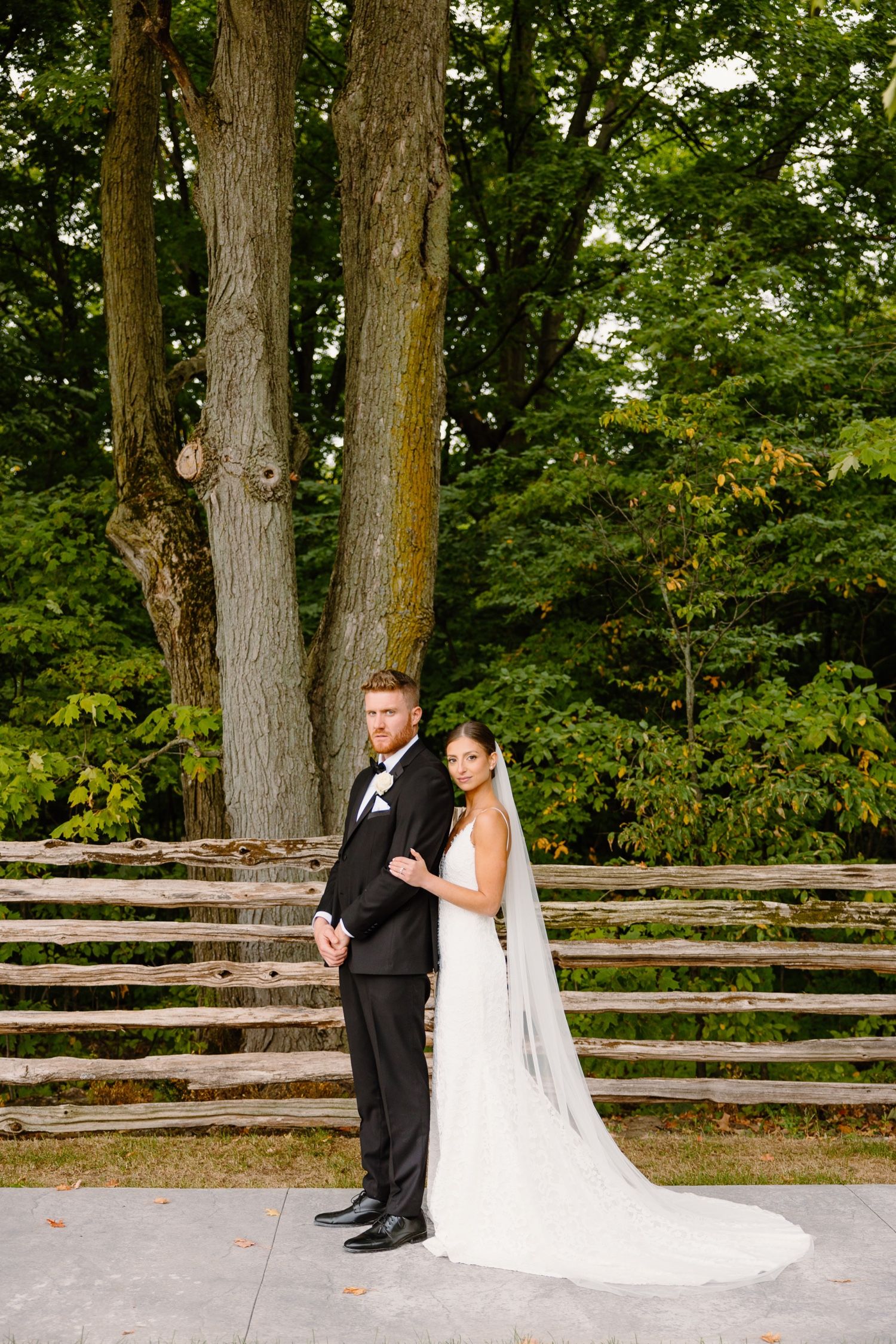 A wedding couple poses in front of rustic wooden fencing and tall trees in an outdoor setting.