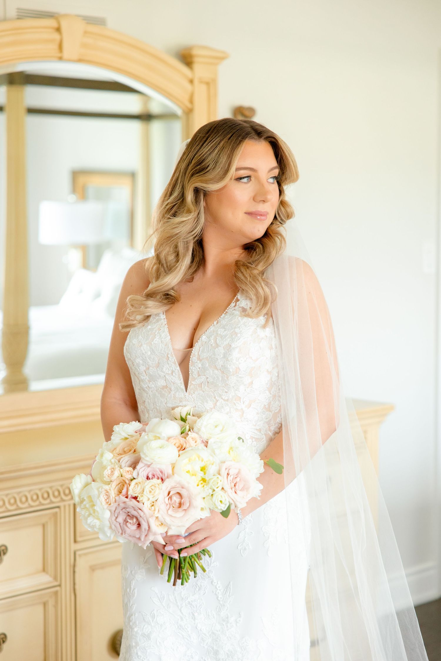 Elegant bridal portrait featuring a lace wedding dress and soft pink and white bouquet in a classic bedroom setting.