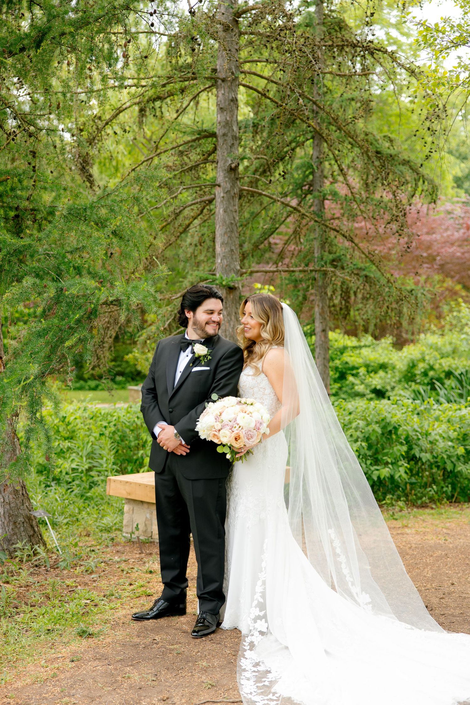 Couple stands together on garden path with flowing cathedral veil and spring foliage backdrop.
