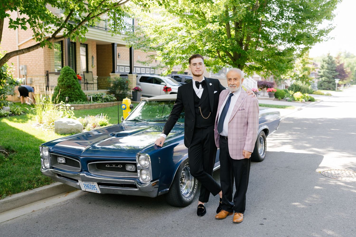 Classic blue car parked on sunny residential street with two people posing beside it.