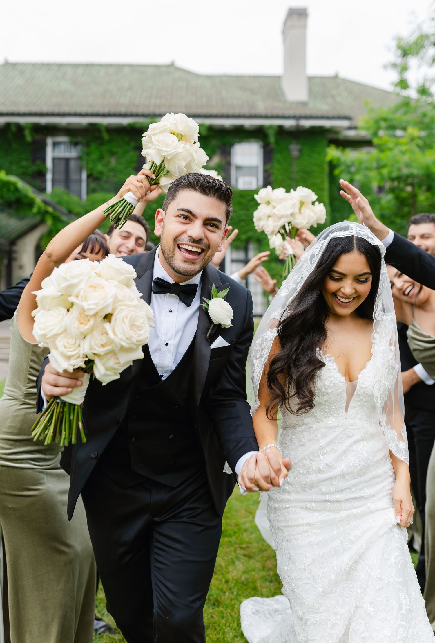 Wedding couple celebrates with their party holding white rose bouquets against ivy-covered building.