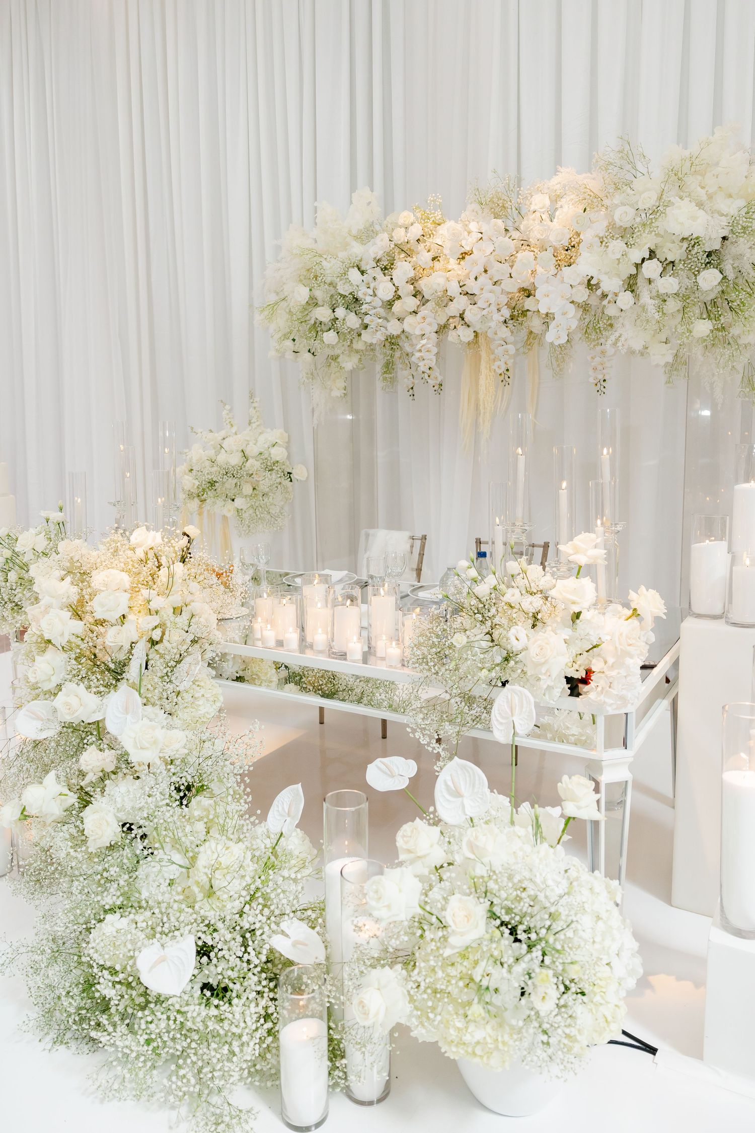 Elegant all-white wedding decor display with cascading flowers and glowing candles on a mirrored table.