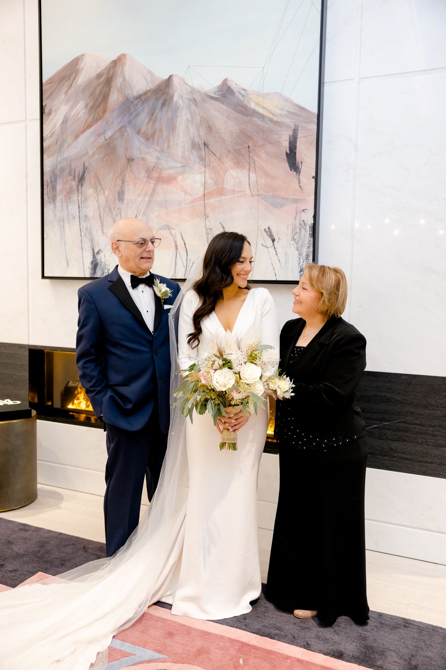 Wedding portrait with bride in white gown and formal dress guests by mountain artwork and fireplace.