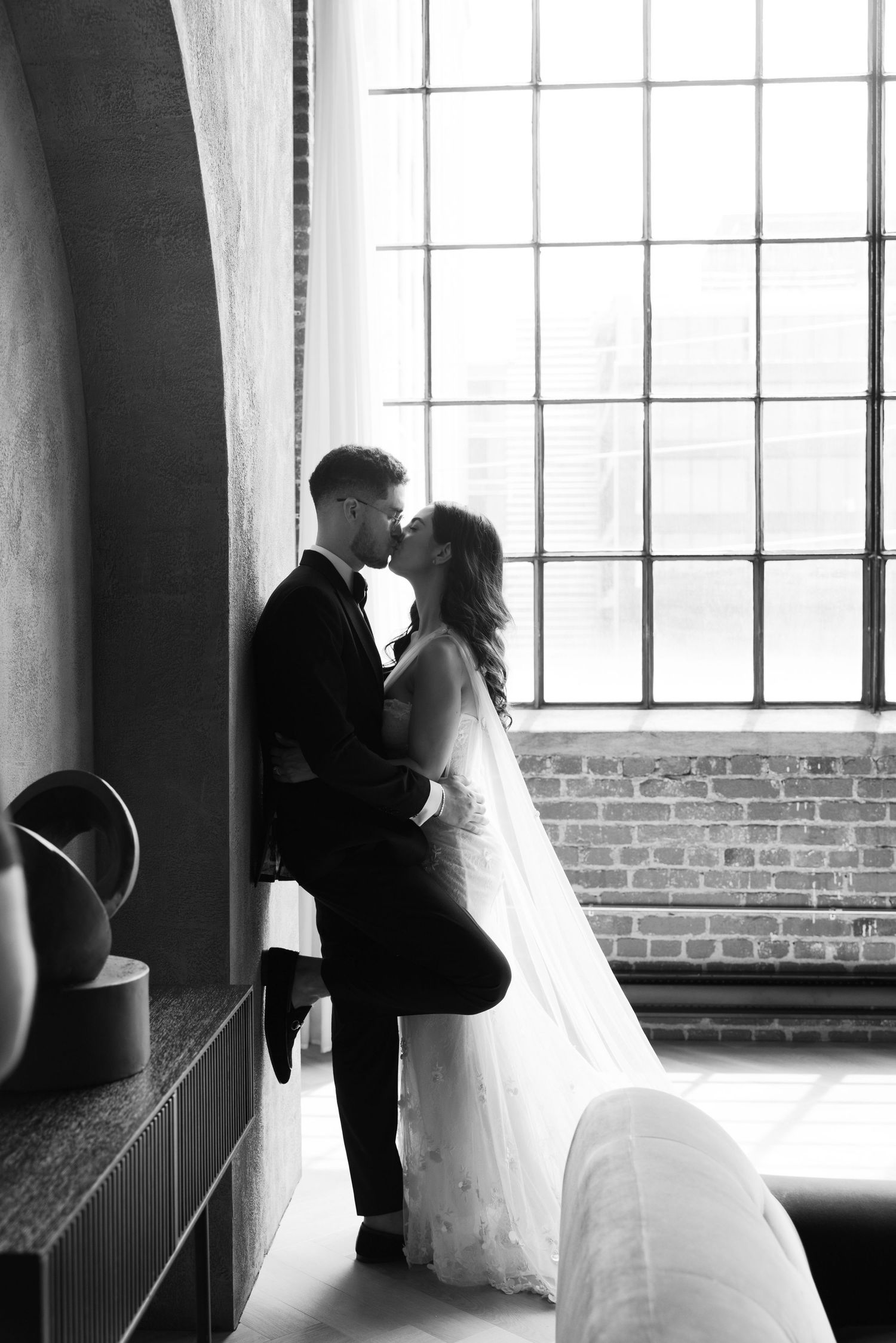 Black and white wedding photo of couple embracing by large industrial window with exposed brick.