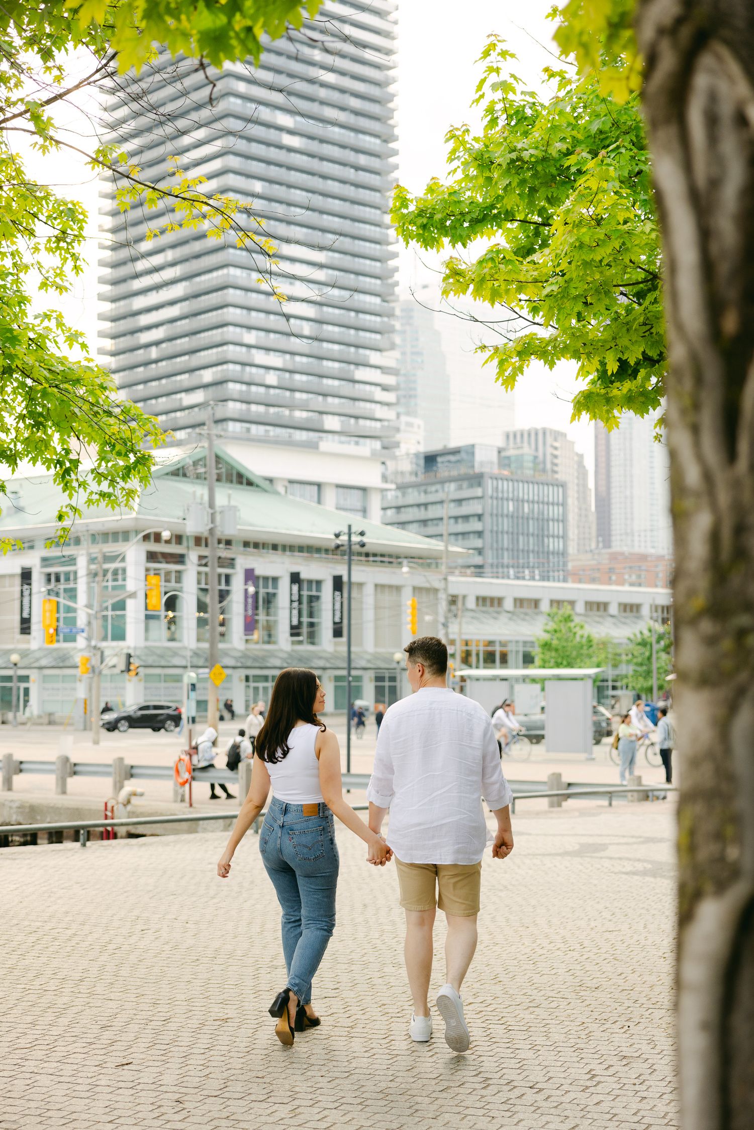 A couple strolls hand in hand through downtown Toronto with modern buildings and green trees in background.