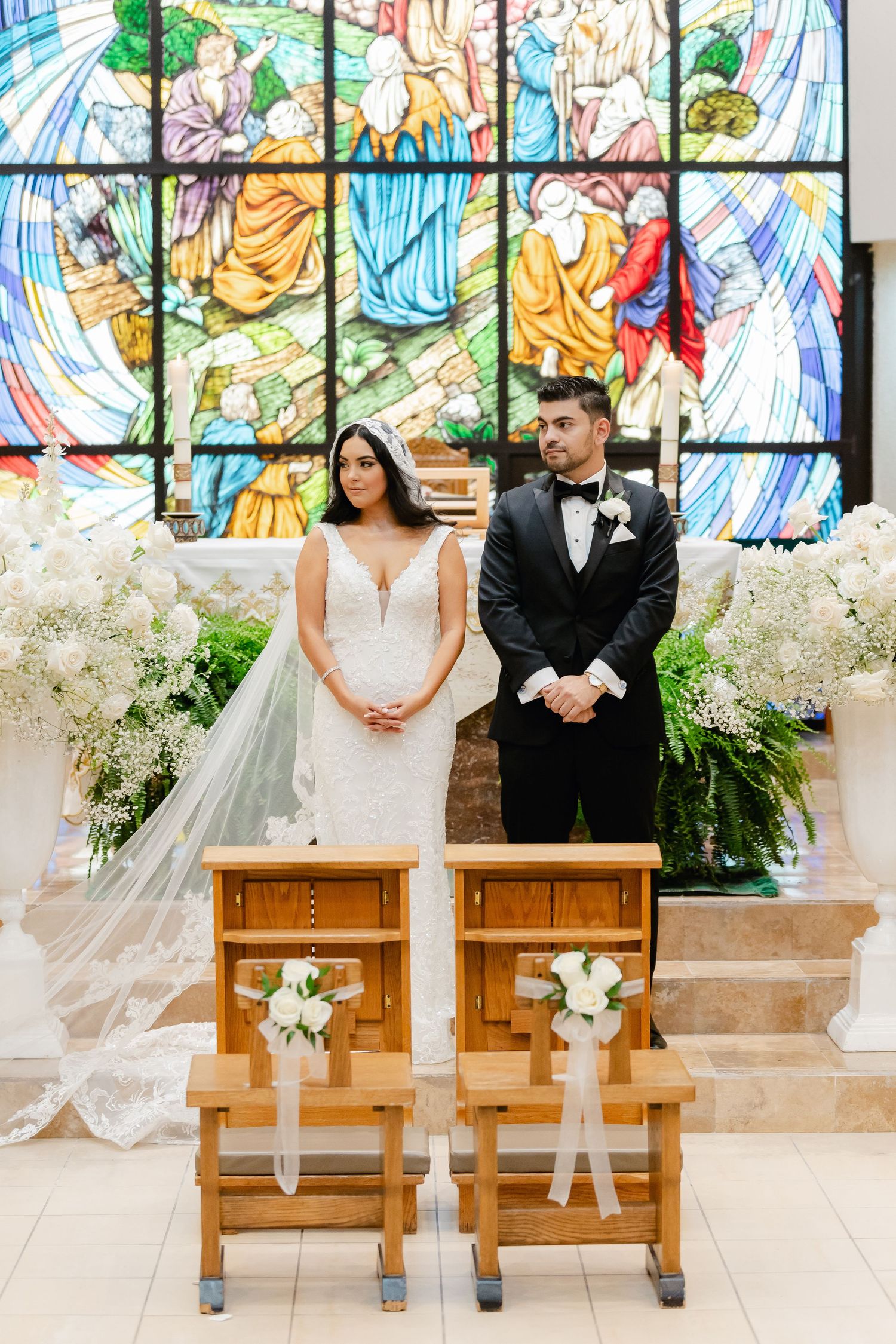 Wedding couple standing before colorful stained glass window with white floral arrangements.