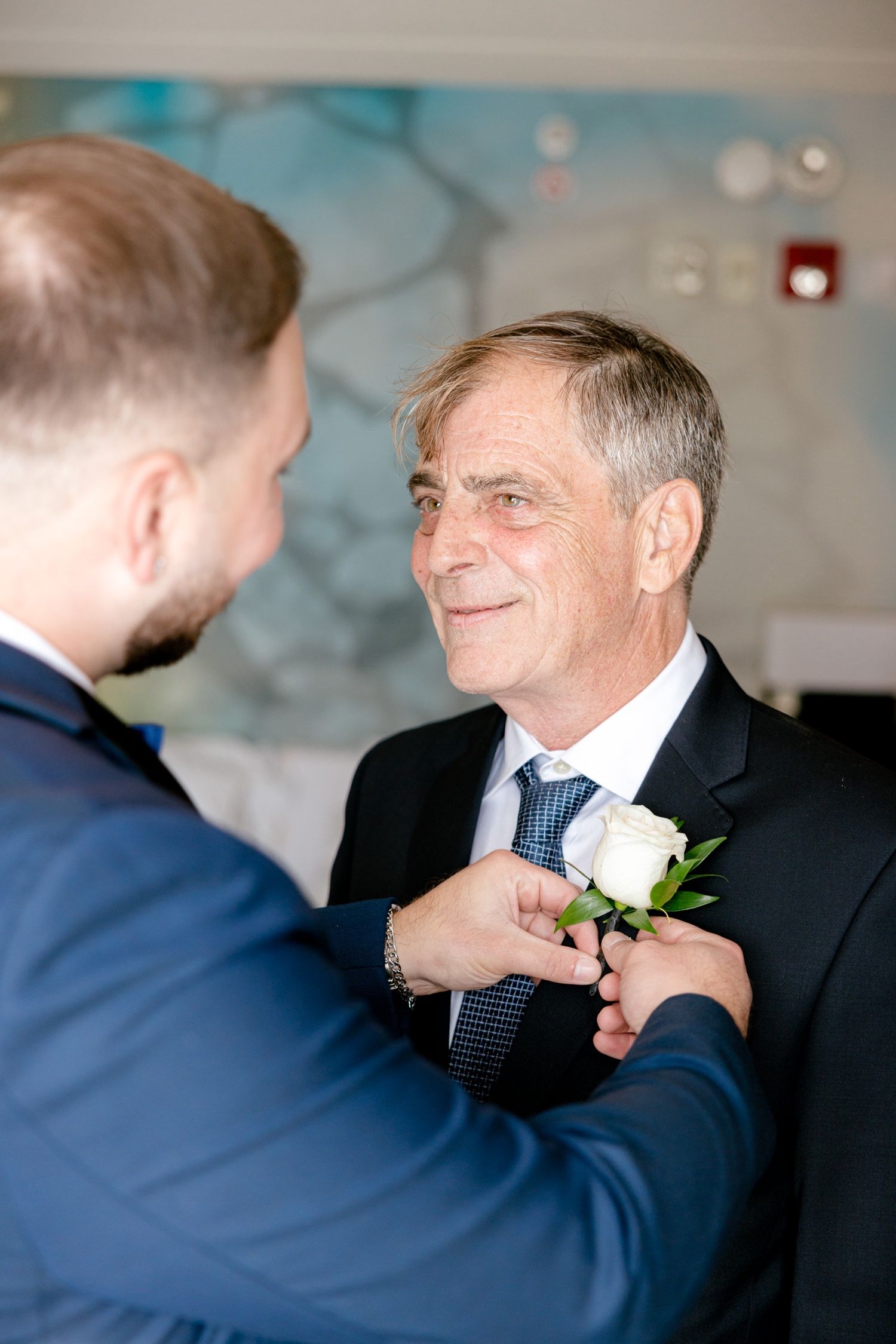 A touching moment as a boutonniere is pinned on a suit lapel against a blue abstract wall backdrop.
