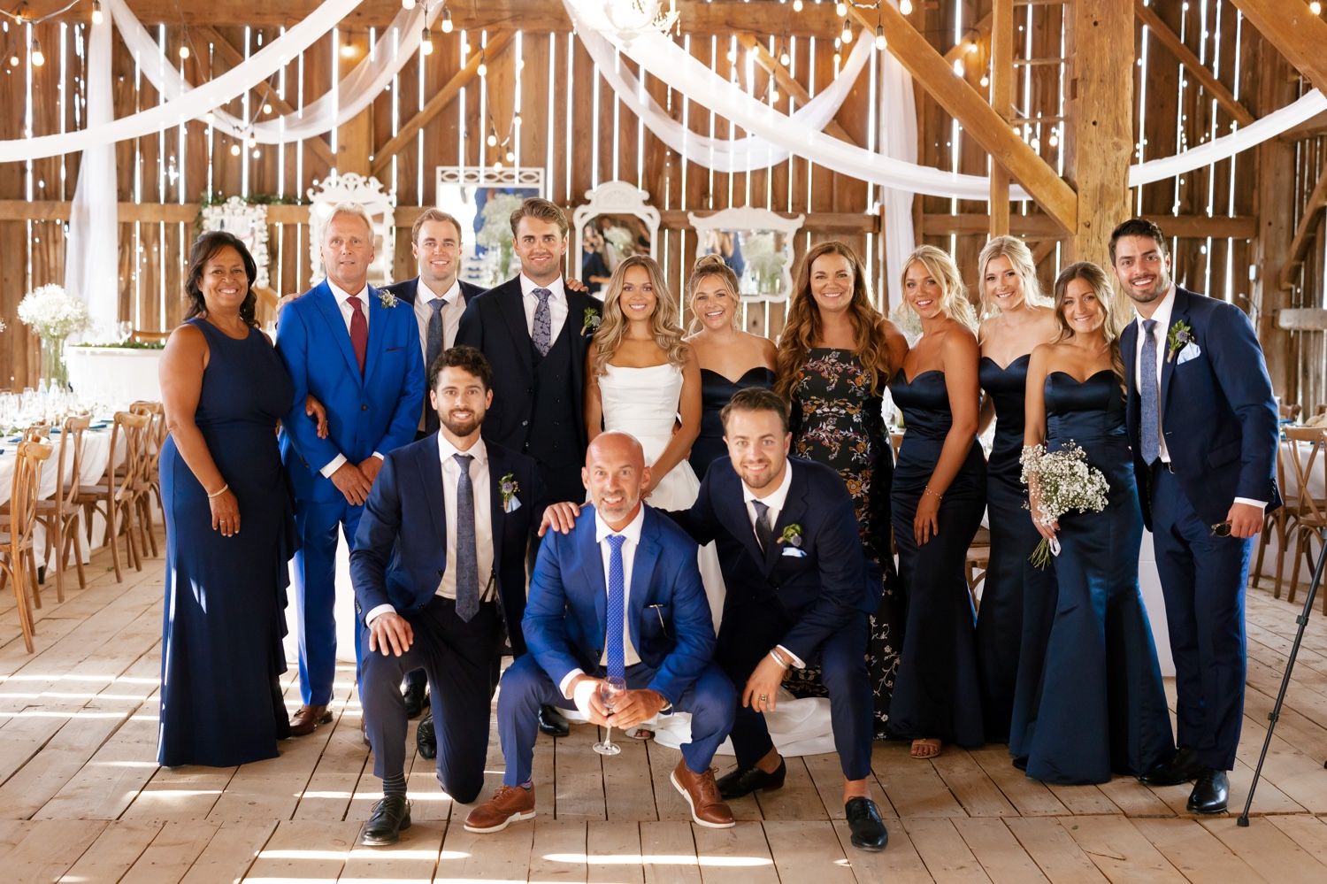 Wedding party gathered in rustic barn venue decorated with white draping and string lights.