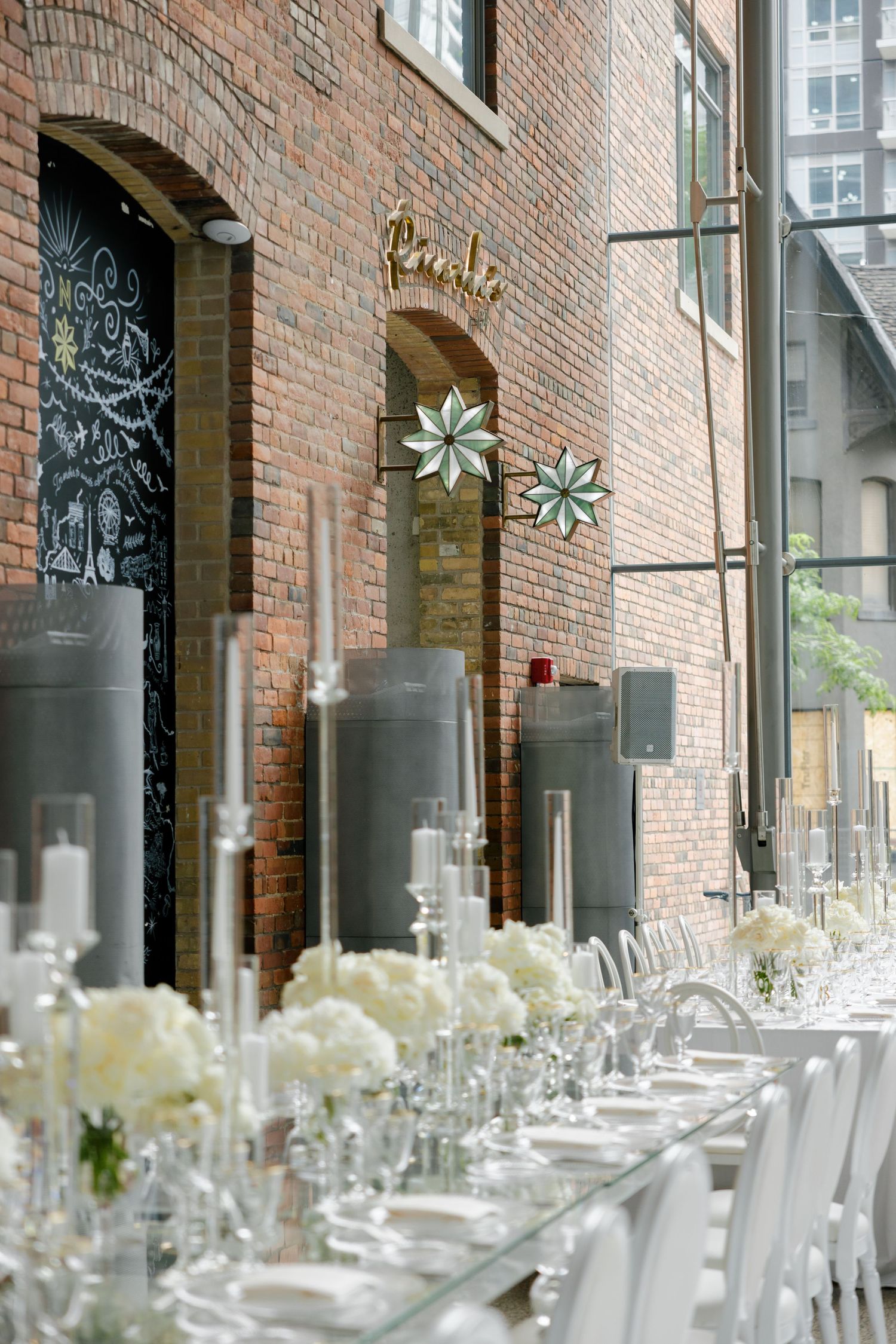 Elegant white table settings with floral centerpieces arranged along exposed brick wall with decorative details.