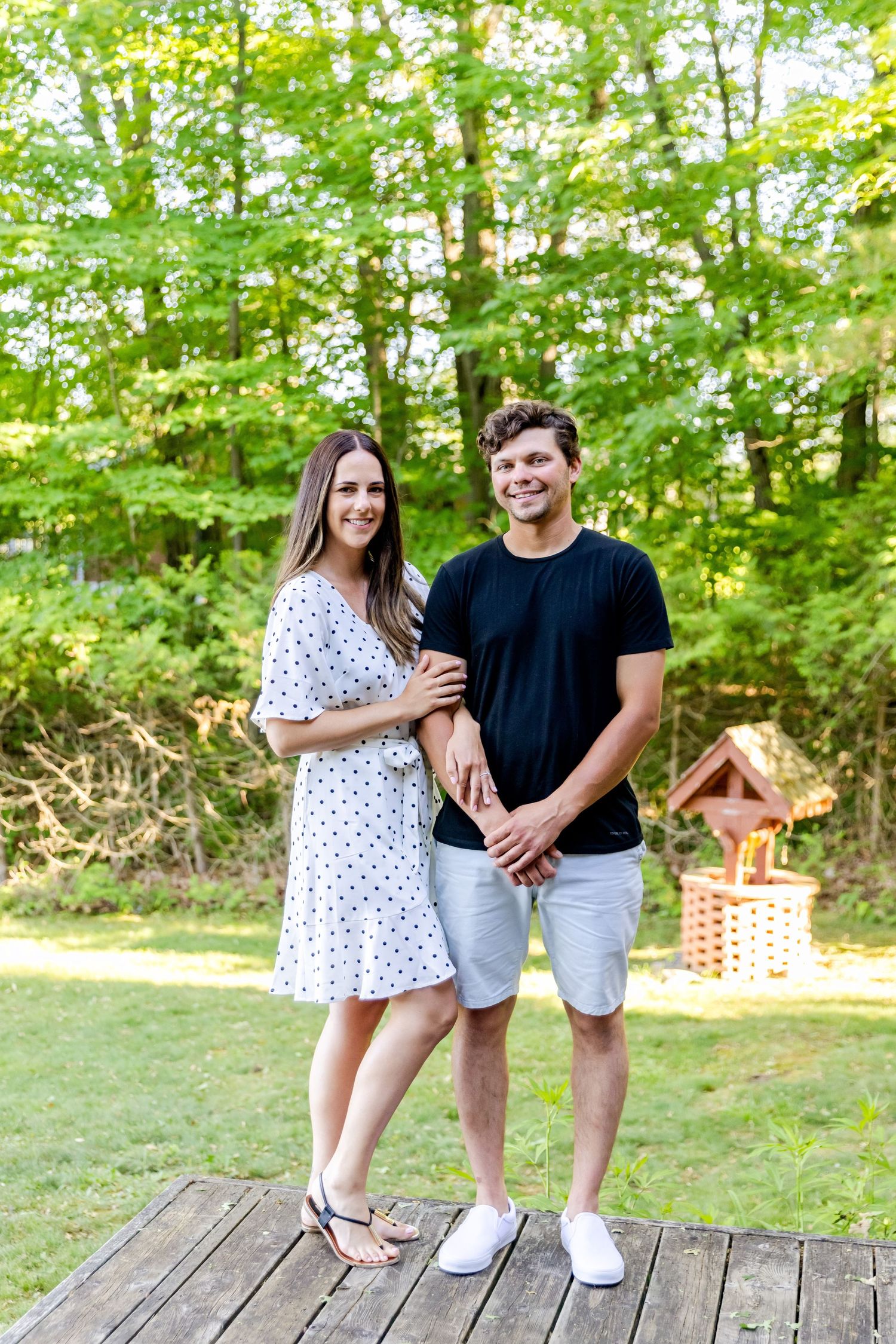 A couple in coordinated black and white outfits poses on a wooden deck with lush greenery in the background.