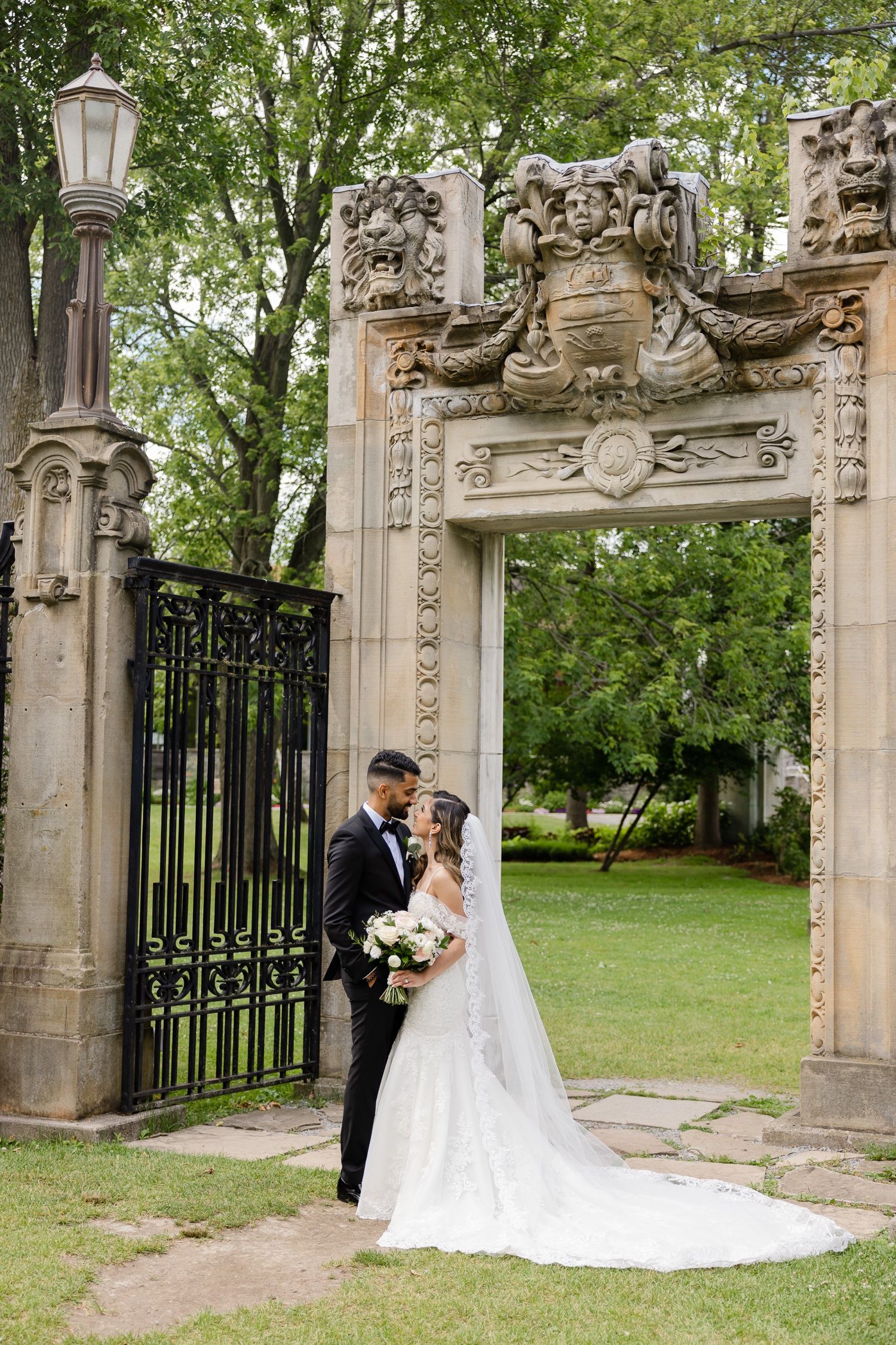 Newlyweds pose beneath an elaborate stone archway with ornate carvings and decorative lantern in a garden setting.
