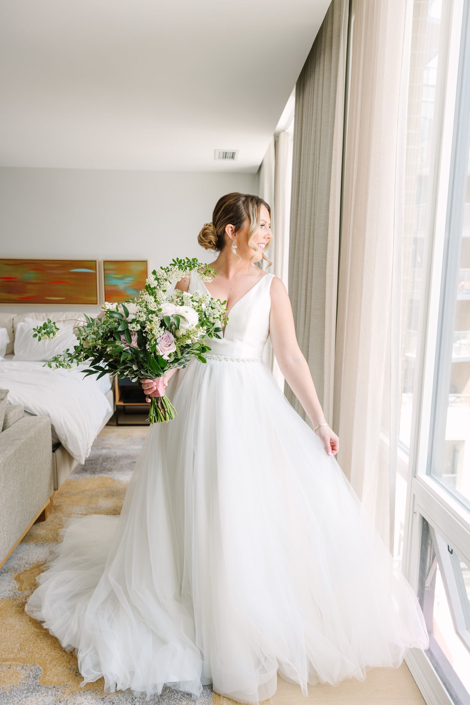 Side profile of bride in flowing white gown holding garden-style bouquet by window.