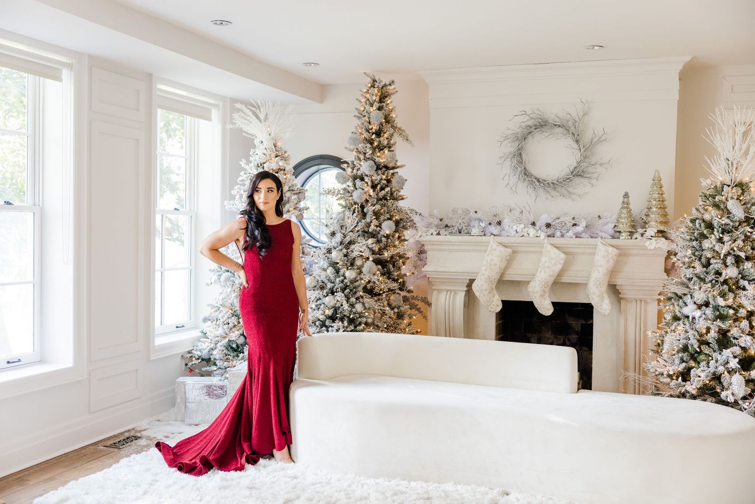 Glamorous holiday photoshoot in red mermaid gown with flocked Christmas trees and white mantel decor.