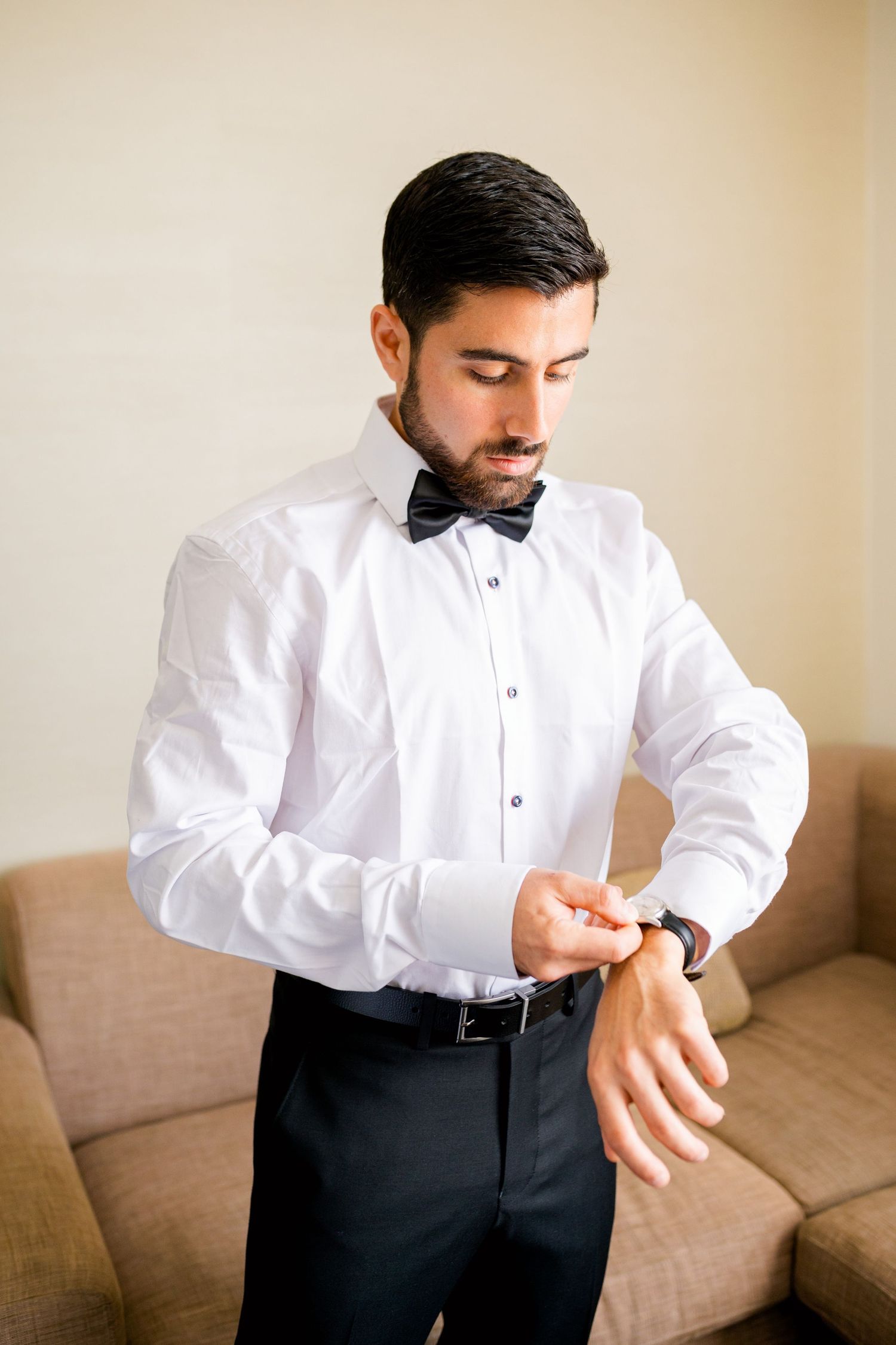 A man in a crisp white shirt and bowtie checks his watch while getting dressed.