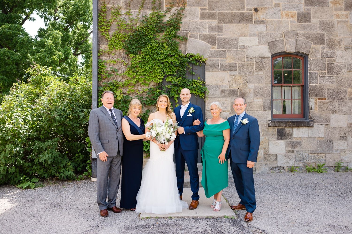 Wedding party poses against stone wall with green foliage and white floral bouquet.