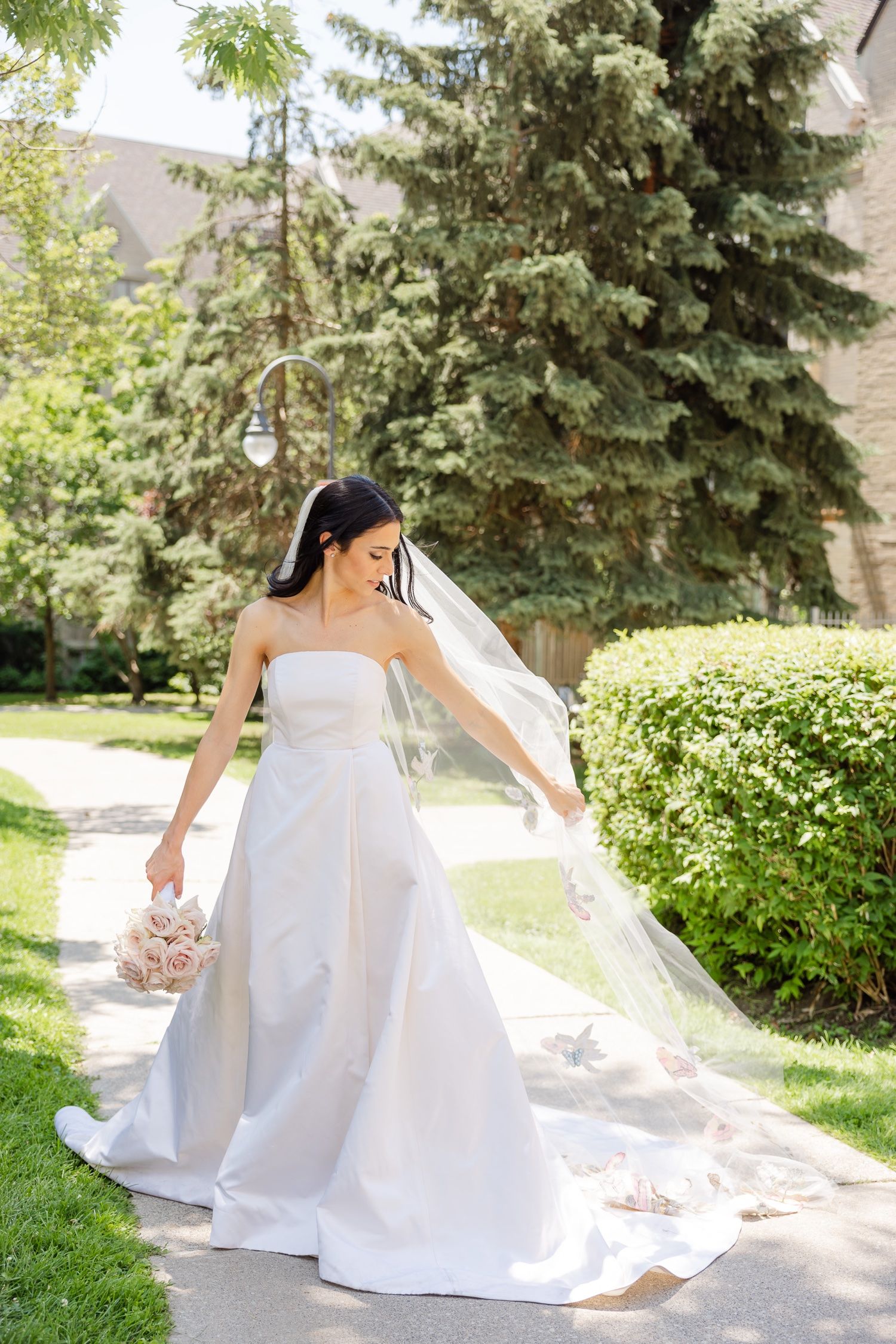 Bride in elegant white wedding gown poses with flowing veil in garden setting.