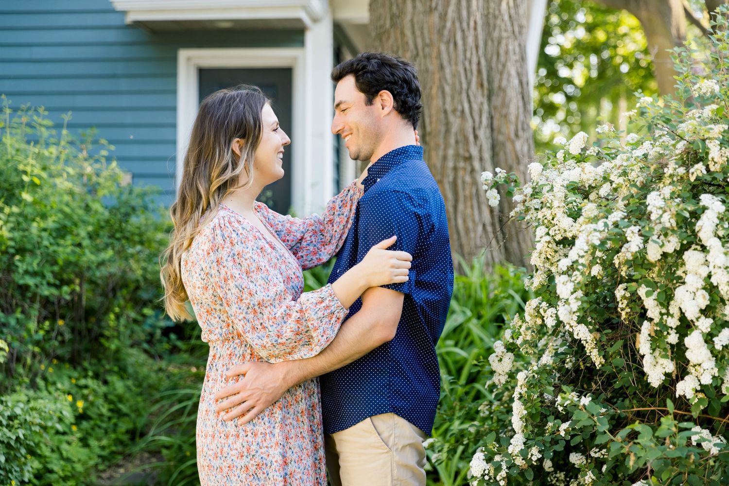 Couple embraces in a garden setting with blooming flowers and blue house in background.