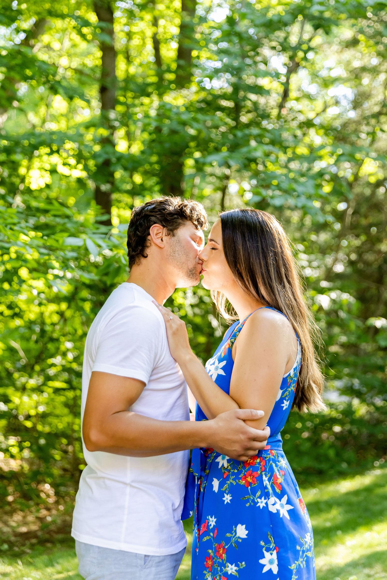 Couple embracing in a sunlit garden wearing white and blue floral dress against lush green foliage background.