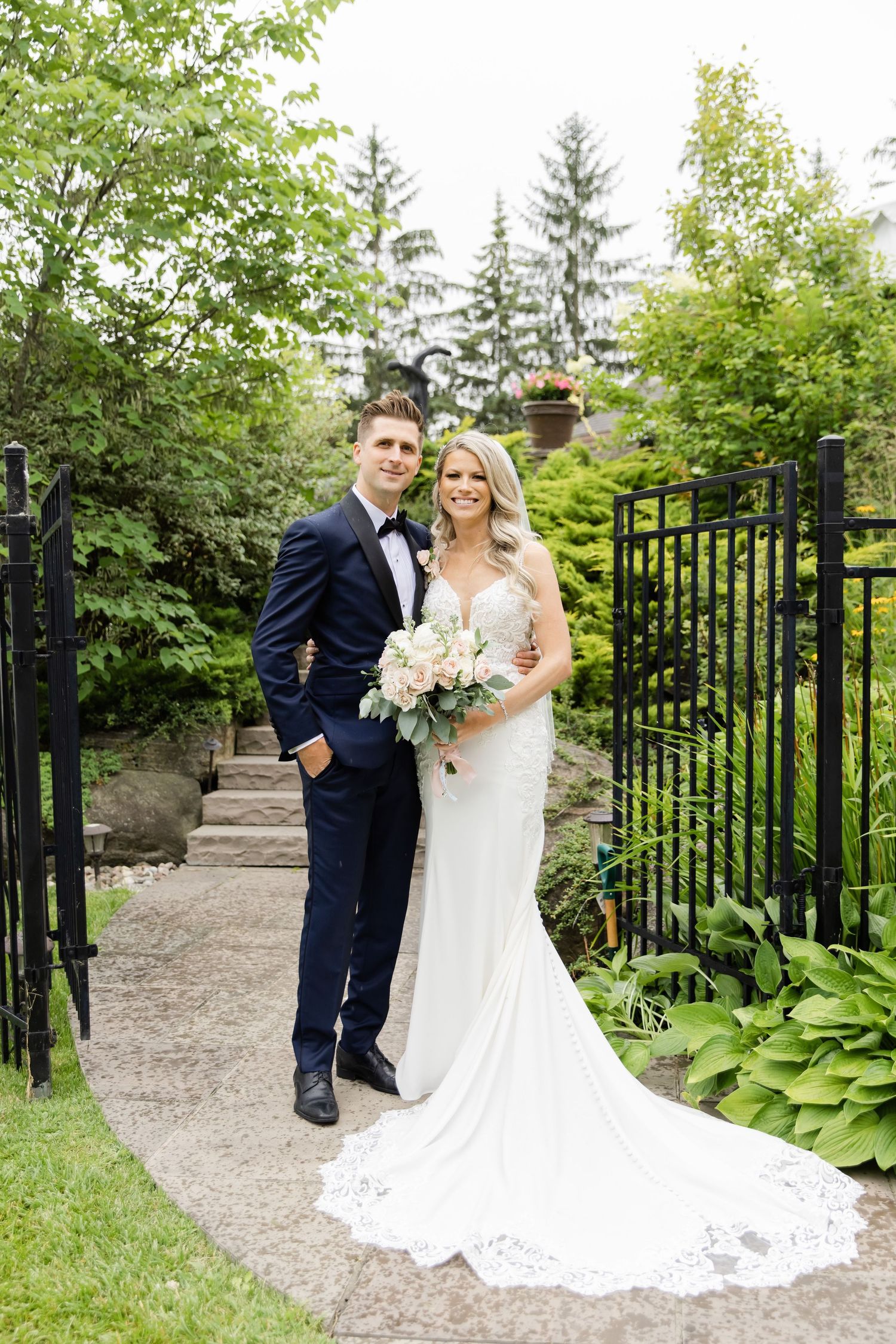 Wedding couple poses on garden path with stone stairs and iron gates.