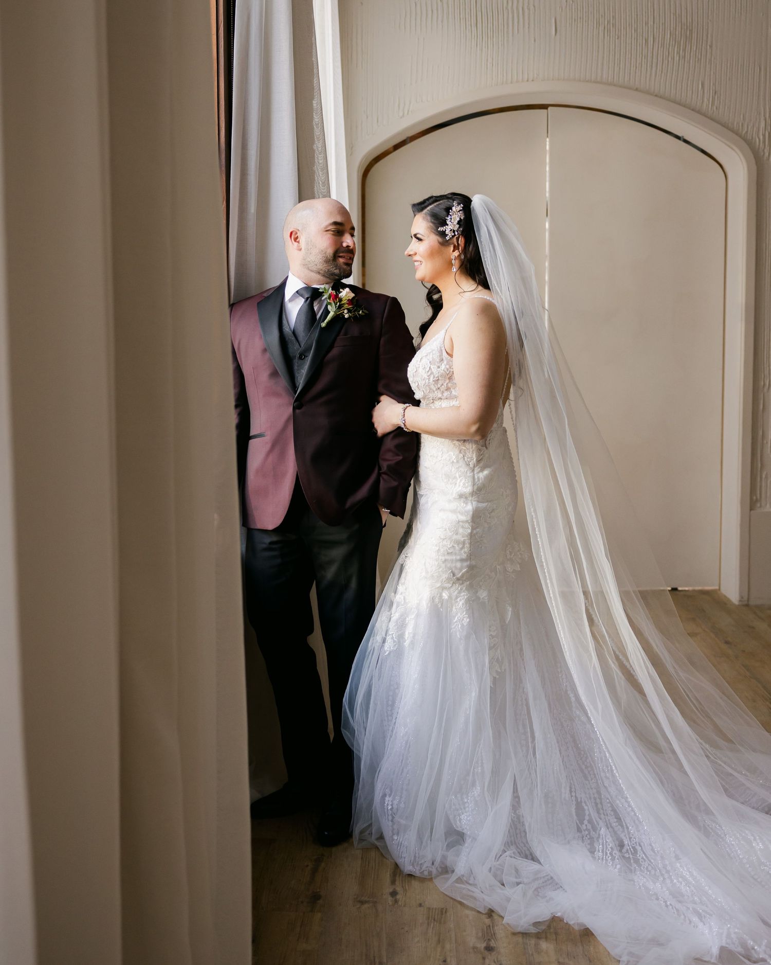 A bride and groom share an intimate moment by a window in a classic indoor setting.