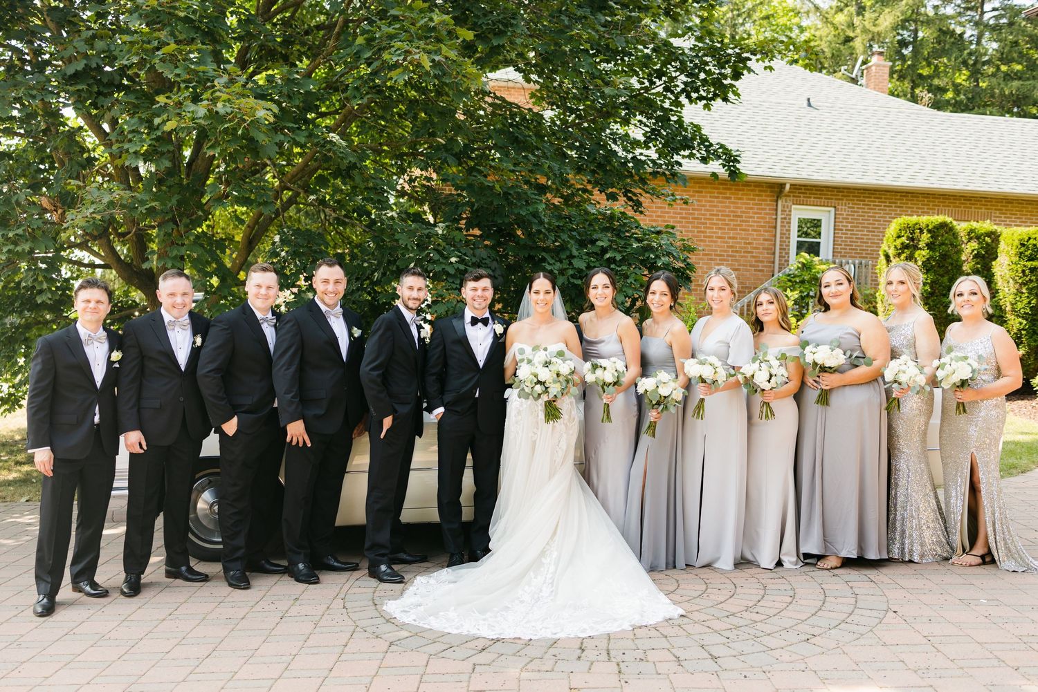 Full wedding party in formal attire posed on brick pavers with bridesmaids in silver and groomsmen in black.
