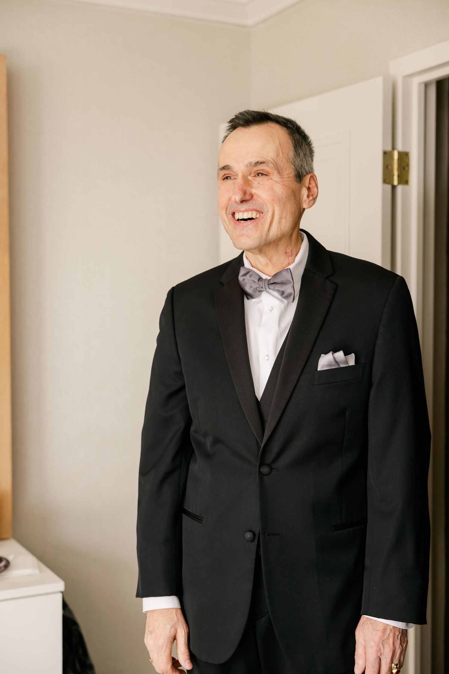 Groom wearing classic black tuxedo and bowtie smiles brightly in getting ready room.