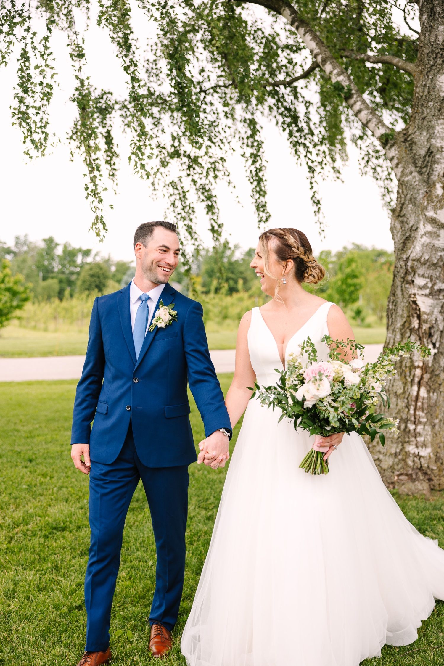 Happy couple holds hands while sharing loving glances under willow tree branches.