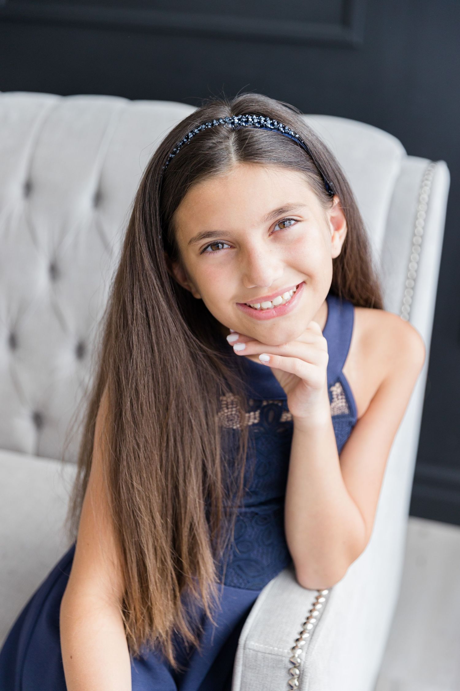Portrait of a young girl in a navy blue dress seated in a white tufted chair with long brown hair.