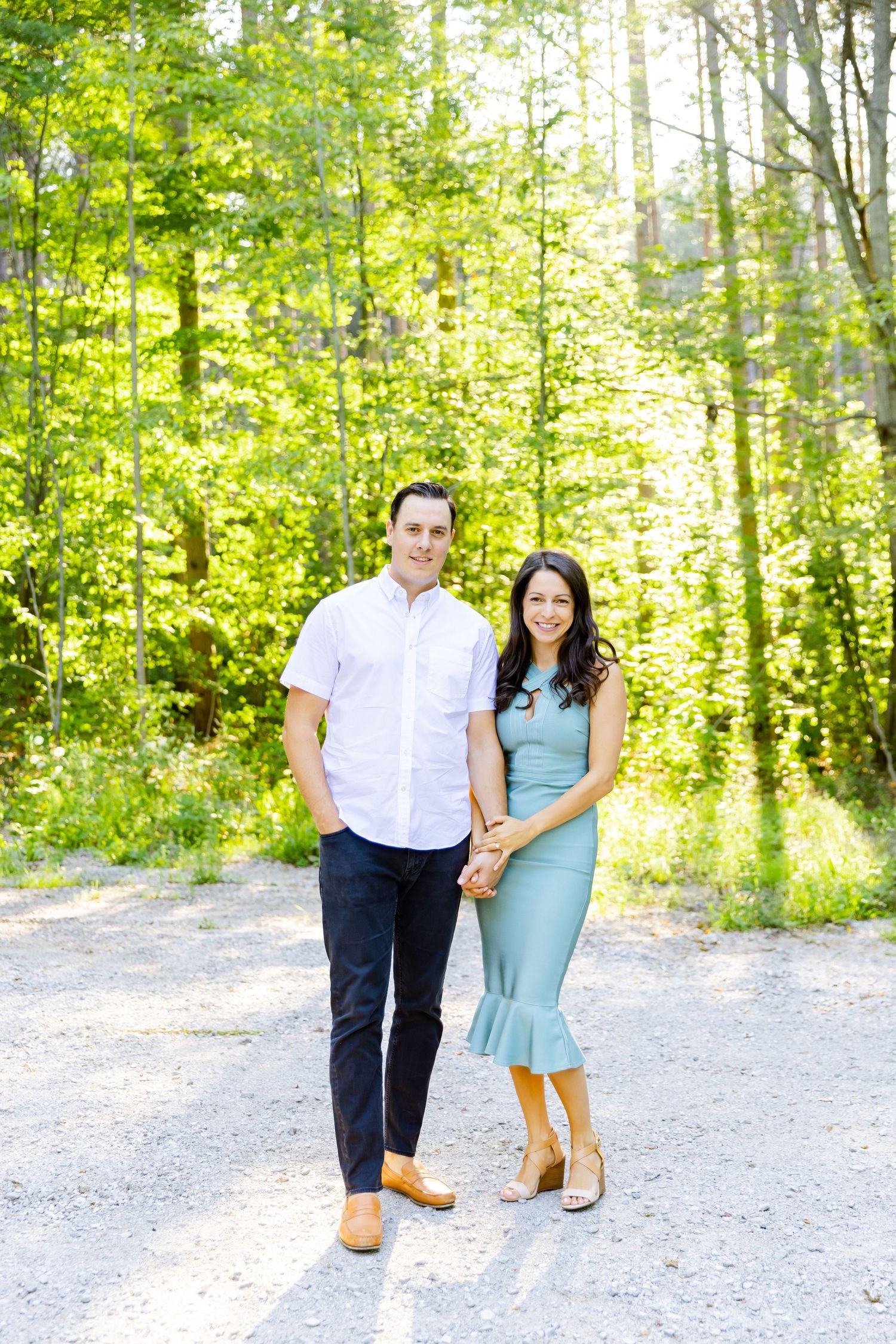 Couple in white shirt and blue dress standing together on sunny forest path.