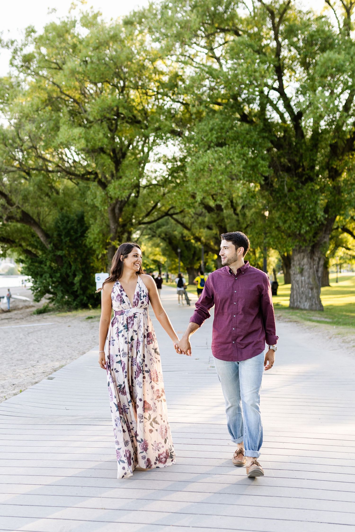 A couple walks hand in hand on a wooden boardwalk under large green trees.