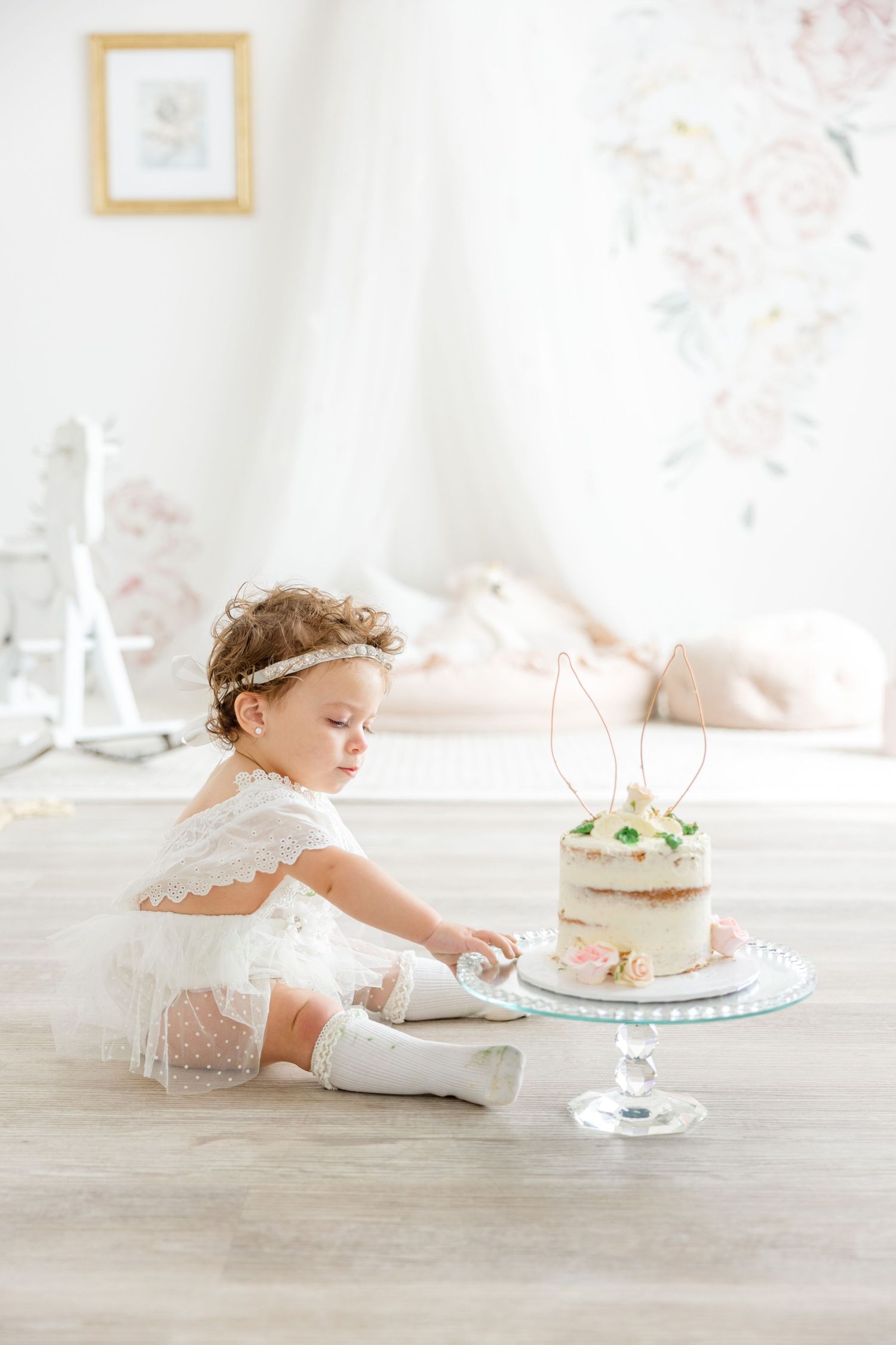 A small child in white lace sits on hardwood floor reaching for a delicate cake with bunny topper.