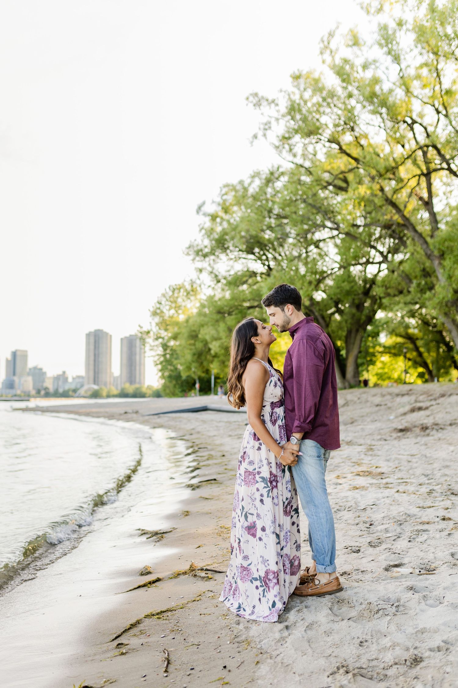 Intimate couple moment captured on sandy beach with city skyline in soft focus background.