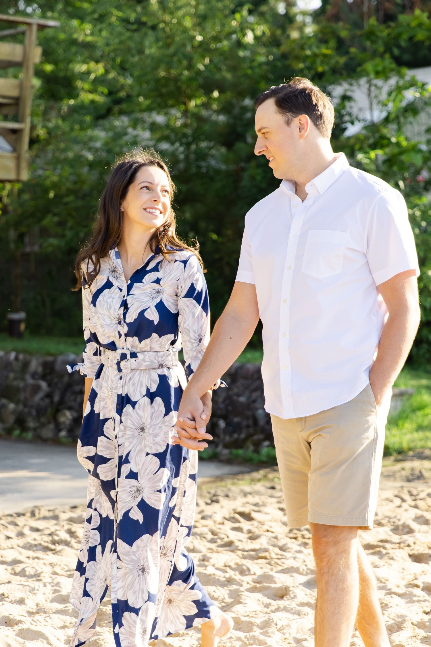 A couple strolls hand in hand outdoors, with her in a blue floral maxi dress and him in a white shirt and khaki shorts.
