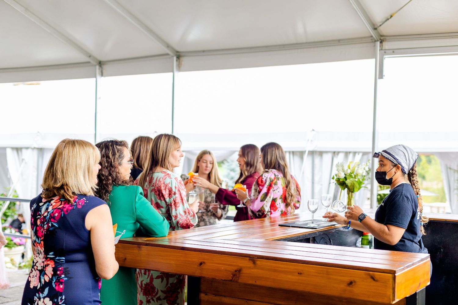 Guests gather at a wooden bar counter under a white tent during an outdoor event.