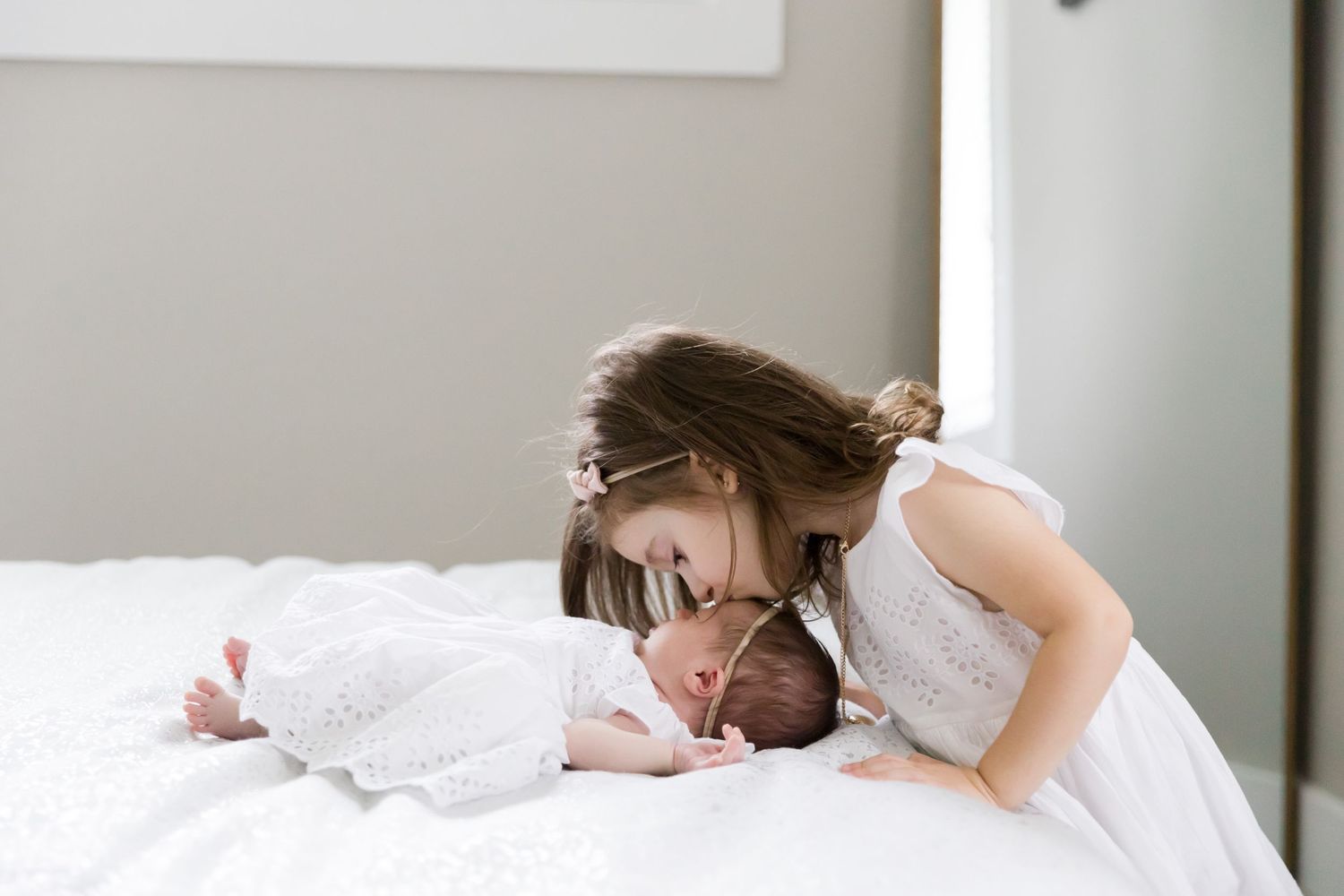 Child giving sleeping baby a gentle kiss on white bedding.
